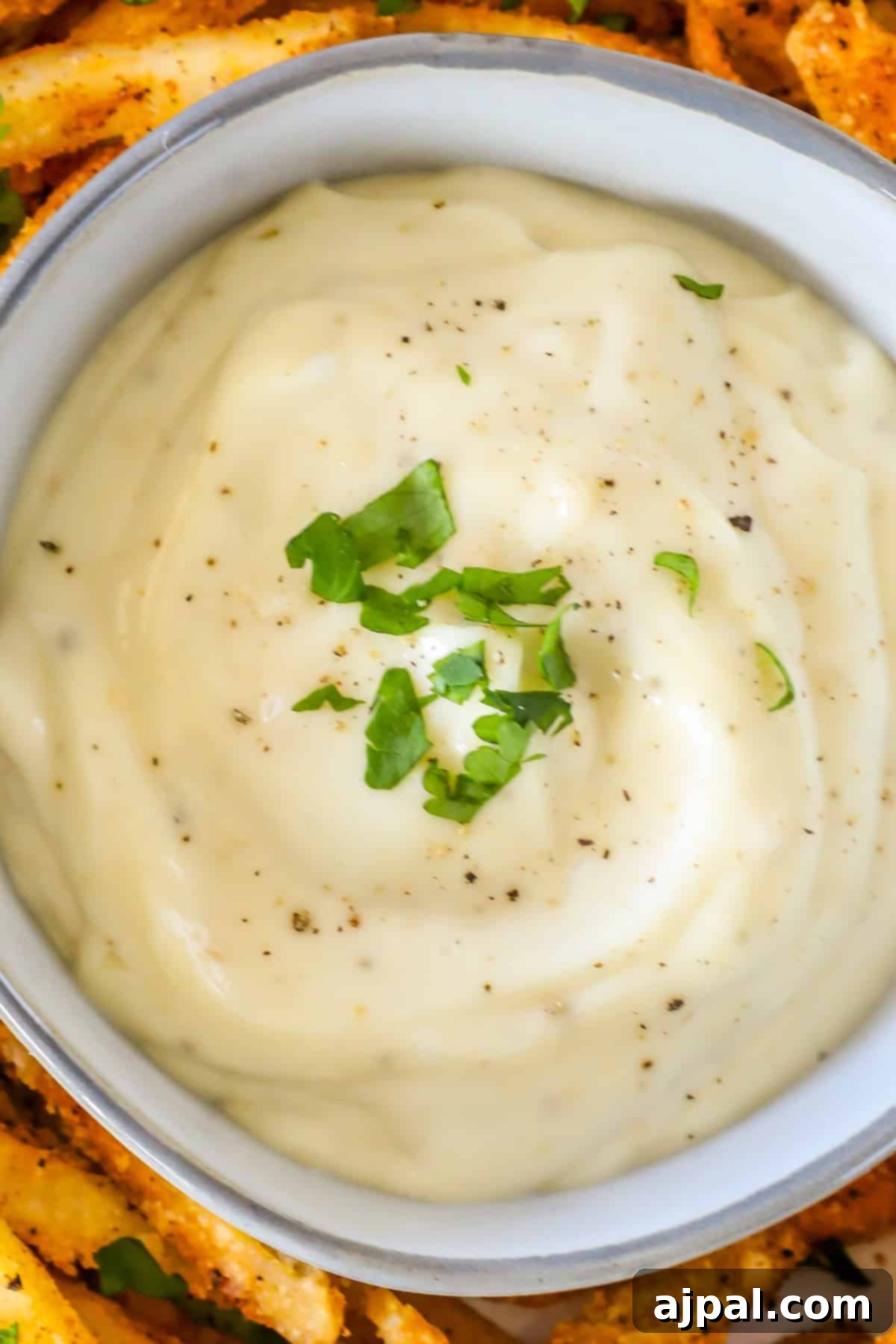 Close-up of a bowl of creamy lemon garlic aioli garnished with chopped green herbs, surrounded by crispy, seasoned potato wedges.