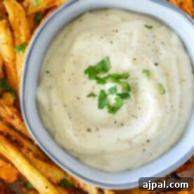 A bowl of creamy lemon garlic aioli garnished with black pepper and green herbs sits surrounded by crispy, seasoned French fries.