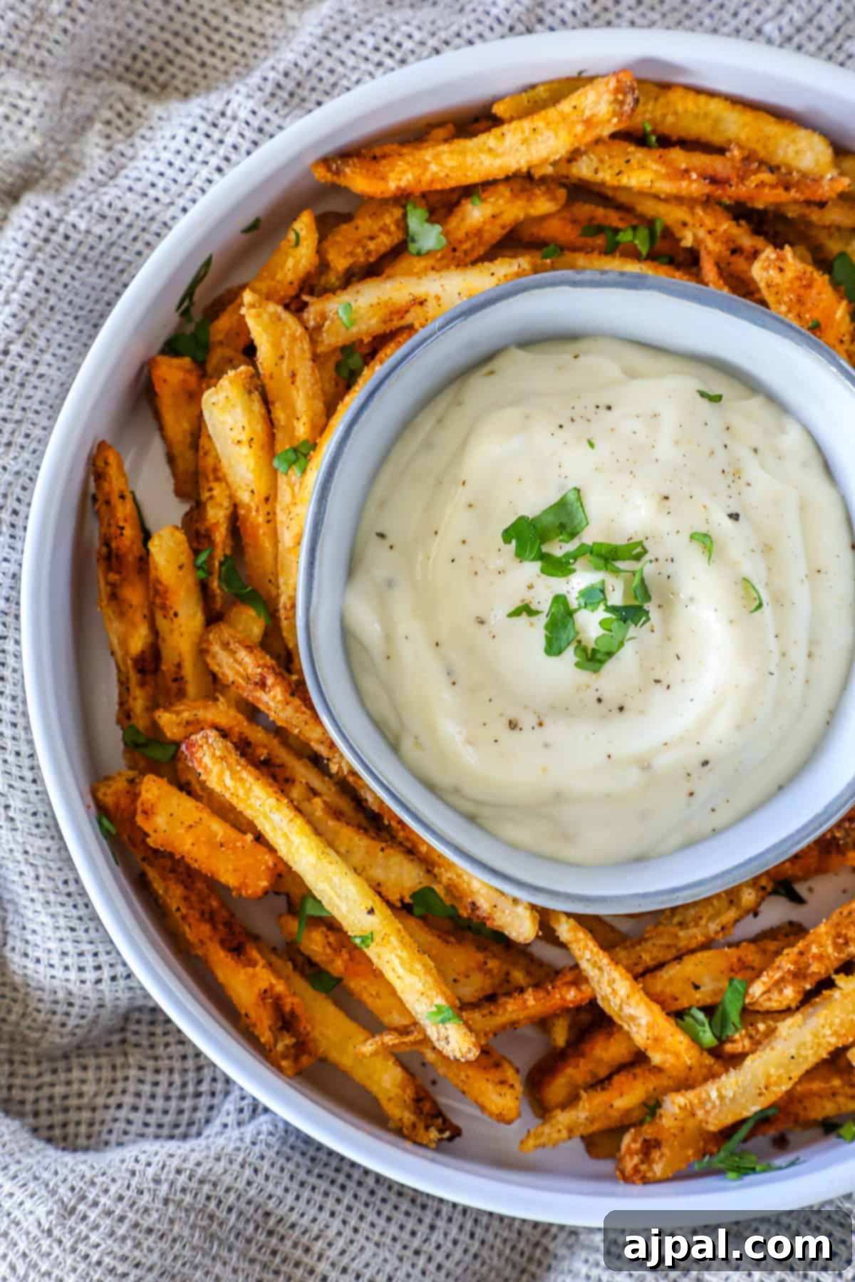 A bowl of crispy, seasoned fries surrounds a smaller dish filled with creamy lemon garlic aioli, garnished with herbs, all set against a textured fabric background.