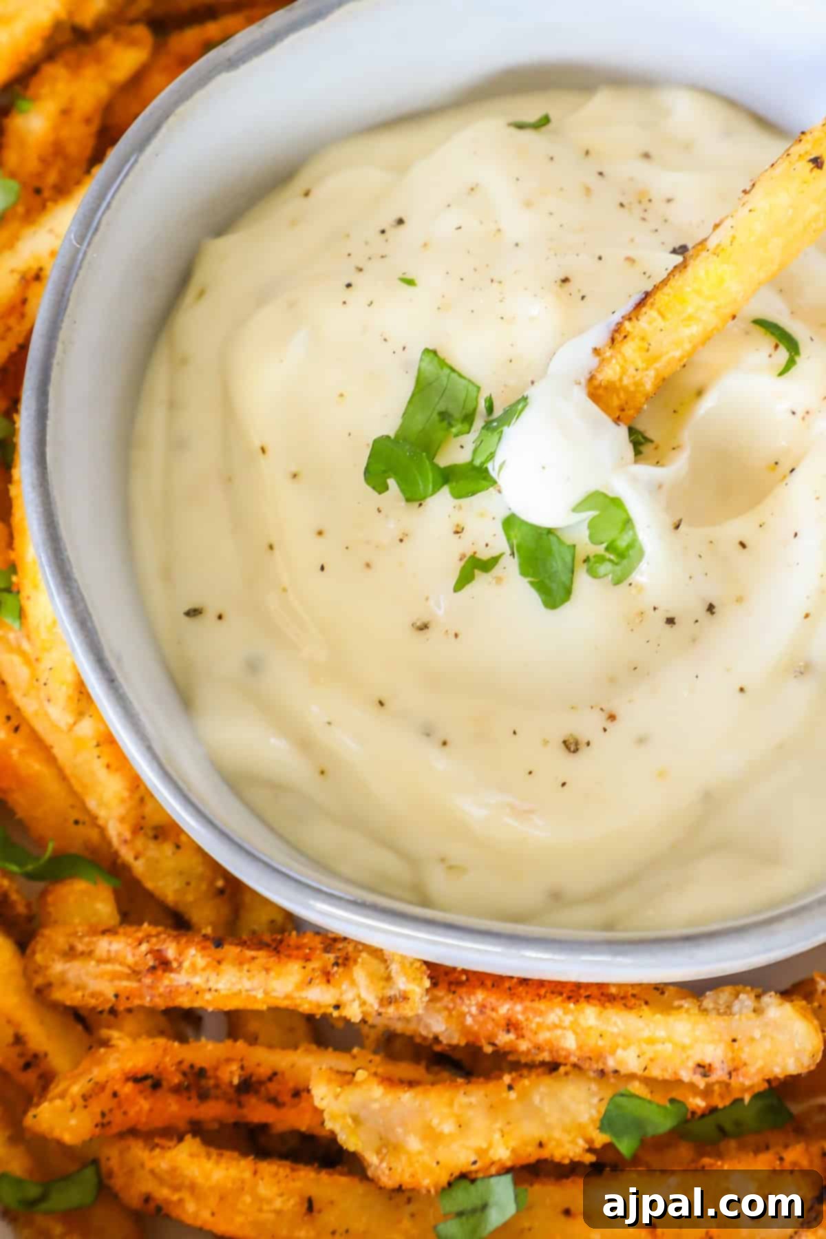 A bowl of creamy lemon garlic aioli topped with herbs, surrounded by seasoned fries. One fry is dipped into the sauce.