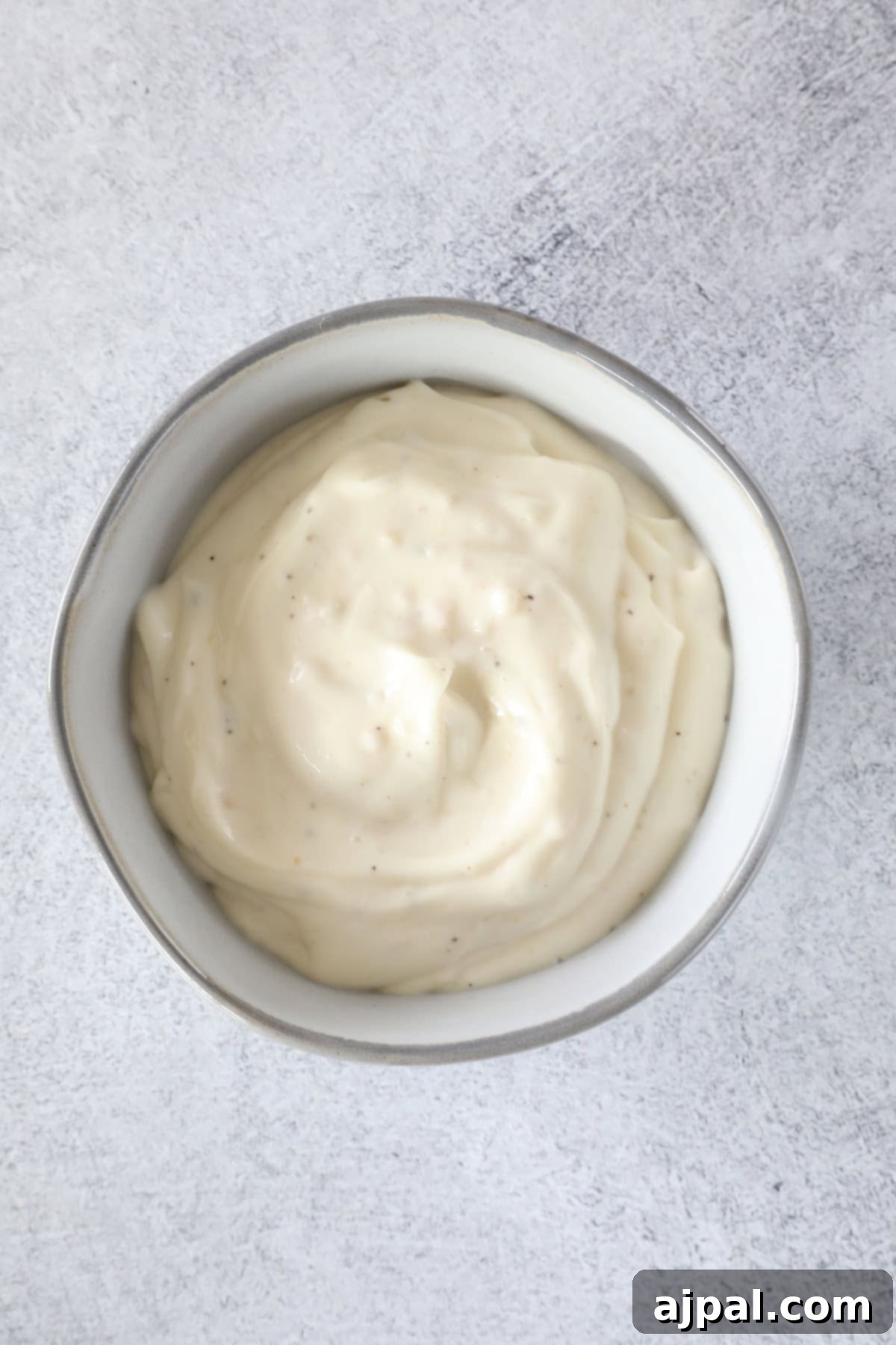 A bowl of creamy, white lemon garlic aioli with a smooth texture sits on a light textured surface.