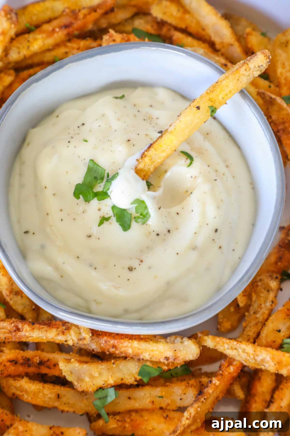 A bowl of creamy lemon garlic aioli garnished with parsley, surrounded by crispy fries seasoned with spices.