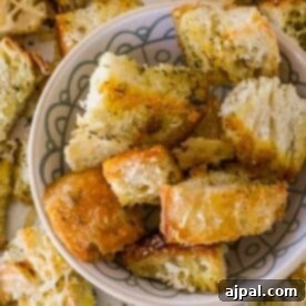 A bowl filled with seasoned sourdough croutons is surrounded by more croutons on a patterned surface.