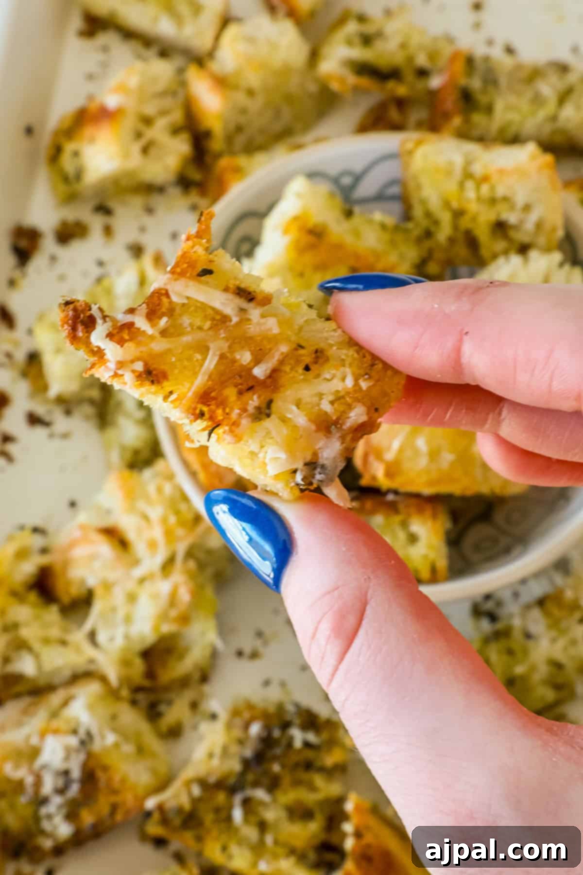 Crispy Sourdough Croutons 3 A person with blue nail polish holds a piece of cheesy bread with herbs, surrounded by more pieces and sourdough croutons on a tray.