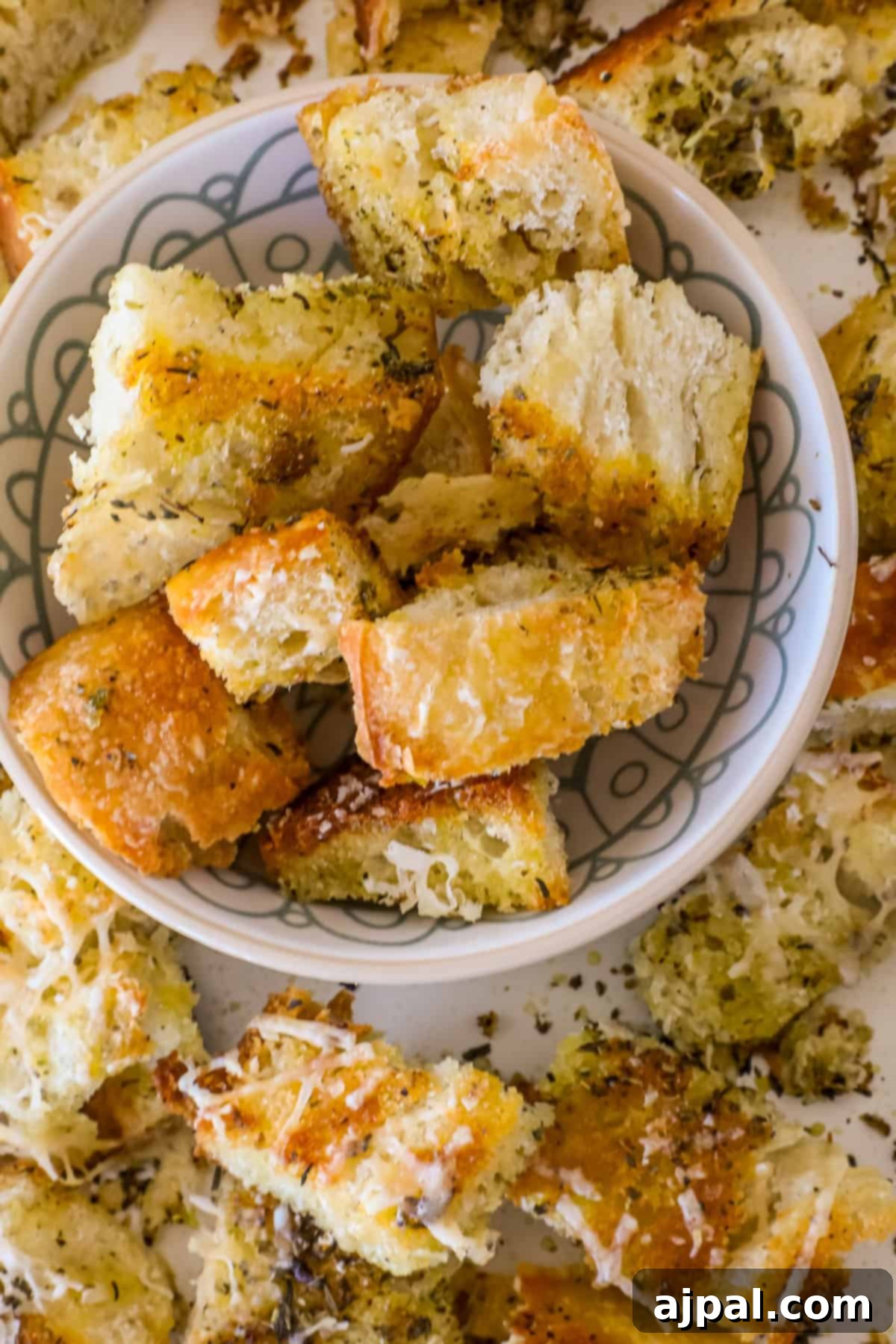 Crispy Sourdough Croutons 7 Sourdough croutons seasoned with herbs and cheese are piled in a bowl and scattered artfully around it on a rustic surface.