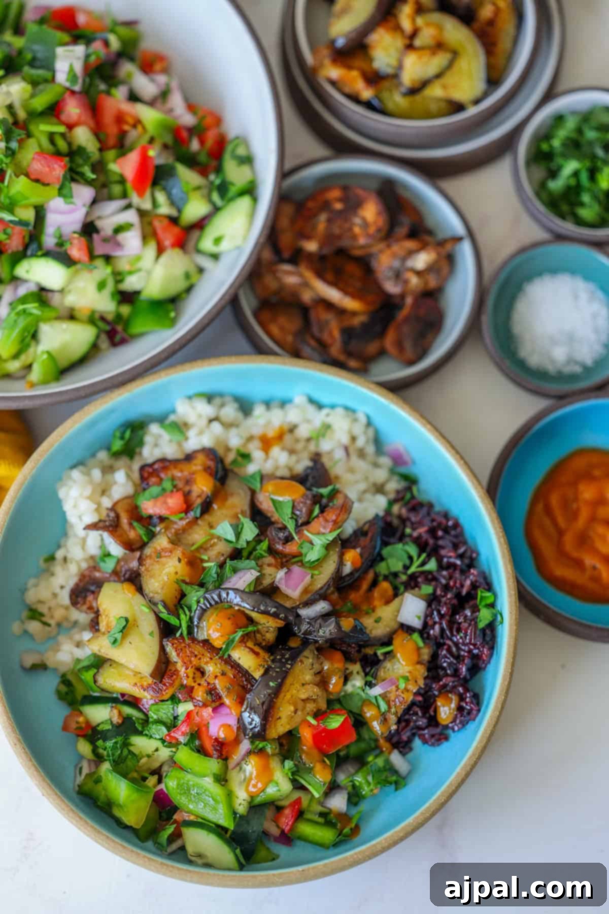 Mediterranean Vegan Sabich Bowls 13 A vegan delight, featuring a bowl of rice topped with grilled vegetables, sits alongside vibrant bowls of salad, eggplant chips, chopped herbs, salt, and sauce on a white surface. This arrangement evokes the essence of Sabich Bowls in its harmonious presentation.