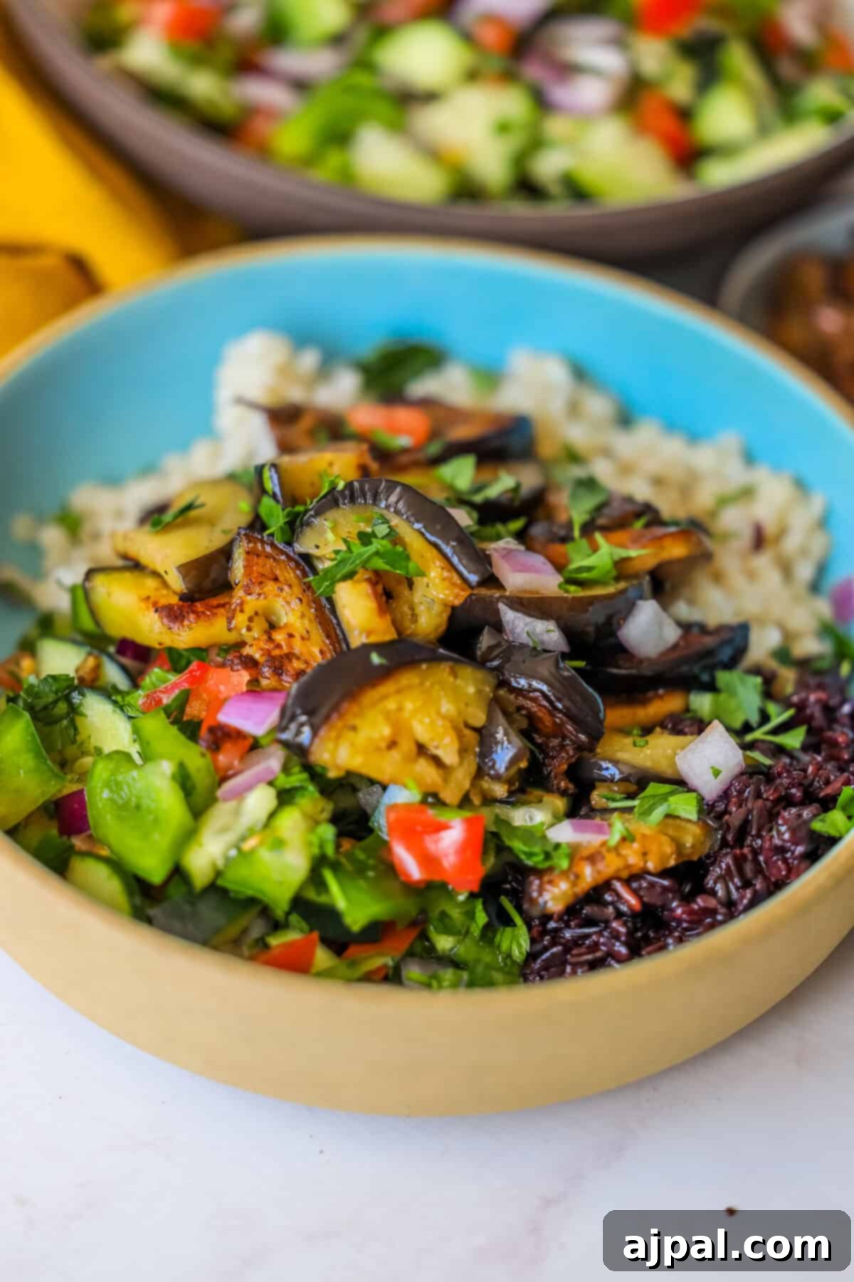 Mediterranean Vegan Sabich Bowls 4 A bowl of mixed grains topped with roasted eggplant, diced tomatoes, cucumbers, and chopped herbs captures the essence of a vegan delight. Another bowl of Sabich-inspired ingredients sits partially visible in the background, enhancing the vibrant scene.
