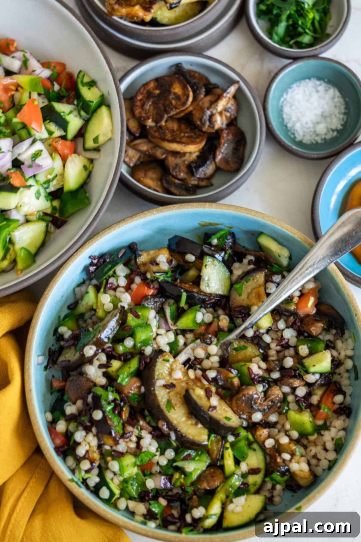 Mediterranean Vegan Sabich Bowls 5 A bowl of vibrant couscous salad with zucchini, bell peppers, and mushrooms sits alongside vegan delights. Surrounding bowls brim with chopped salad, roasted mushrooms, and a touch of salt.