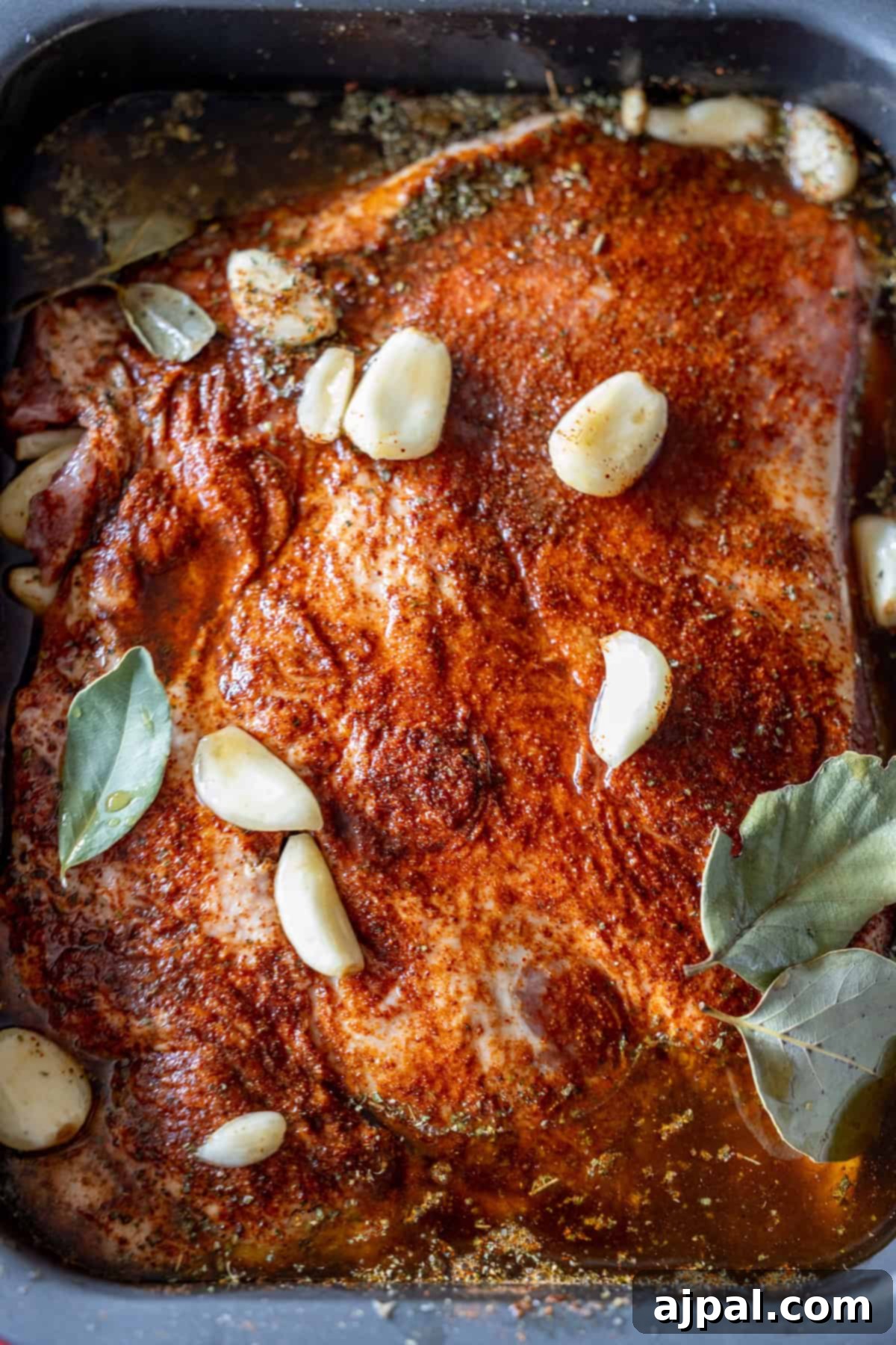 Perfectly Tender Crispy Slow Cooker Carnitas 5 A generously seasoned piece of raw pork, awaiting transformation into flavorful carnitas in a slow cooker, is shown in a pan. It's adorned with fresh garlic cloves, bay leaves (if used for extra aroma), and a medley of spices, all surrounded by a rich marinade.