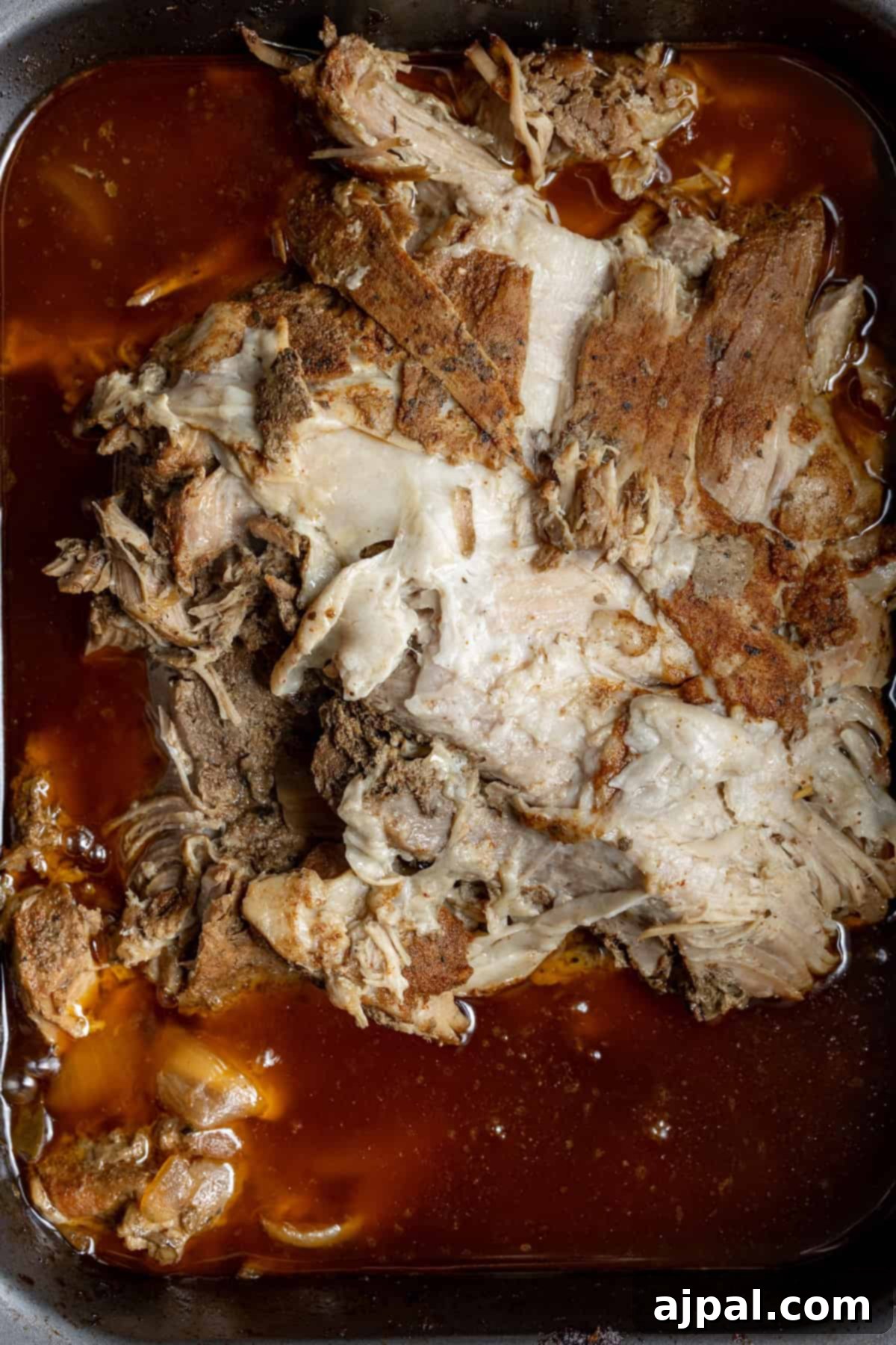 Perfectly Tender Crispy Slow Cooker Carnitas 6 Tender, succulent shredded pork, transformed into Slow Cooker Carnitas, rests in a pan. The meat is immersed in rich, reddish-brown cooking juices, ensuring deep and authentic flavor with every bite.