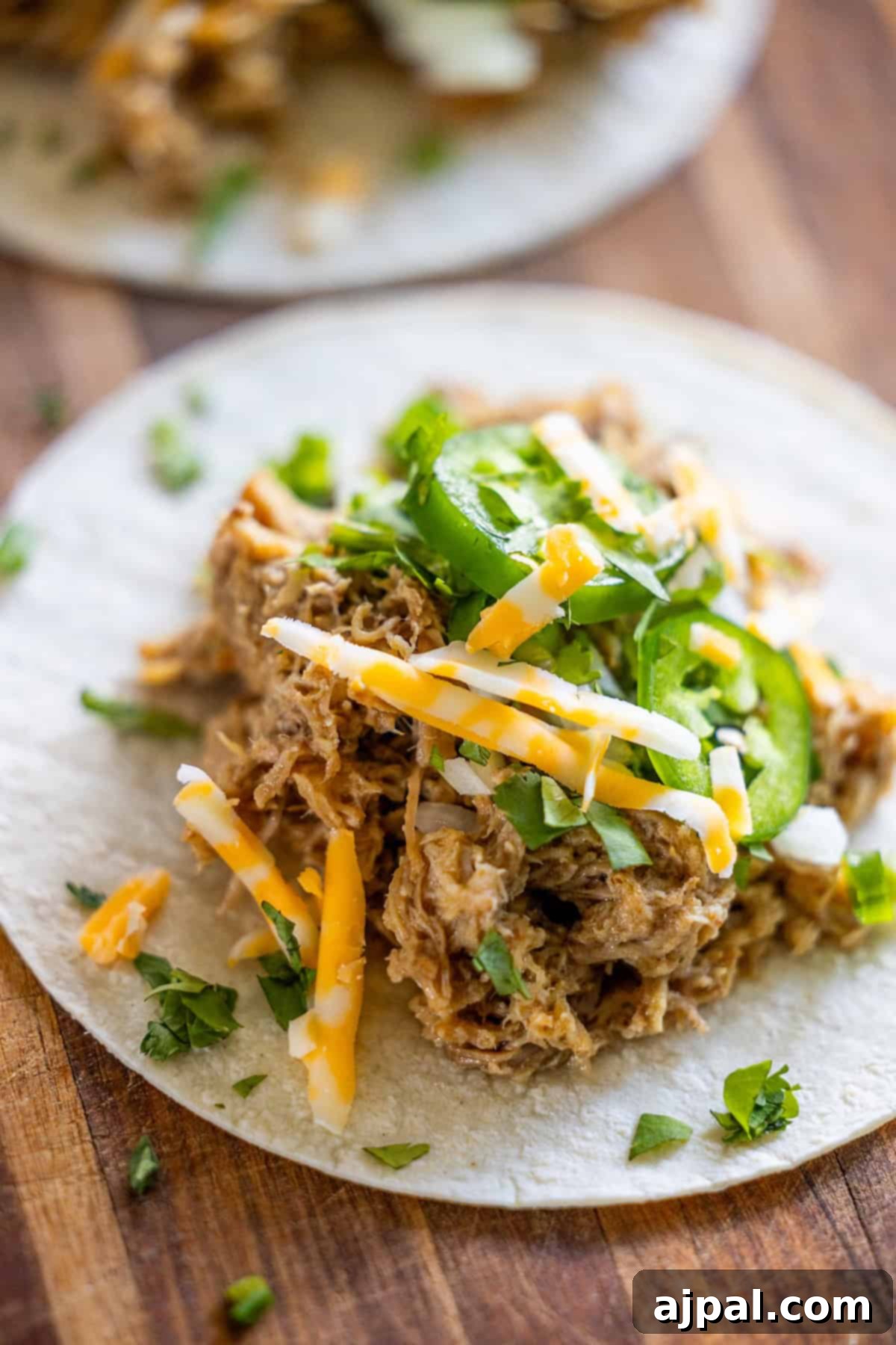 Perfectly Tender Crispy Slow Cooker Carnitas 8 A vibrant flour tortilla topped with succulent slow cooker carnitas, a generous layer of grated cheese, bright jalapeño slices, and a sprinkle of fresh chopped cilantro, all presented on a rustic wooden surface.
