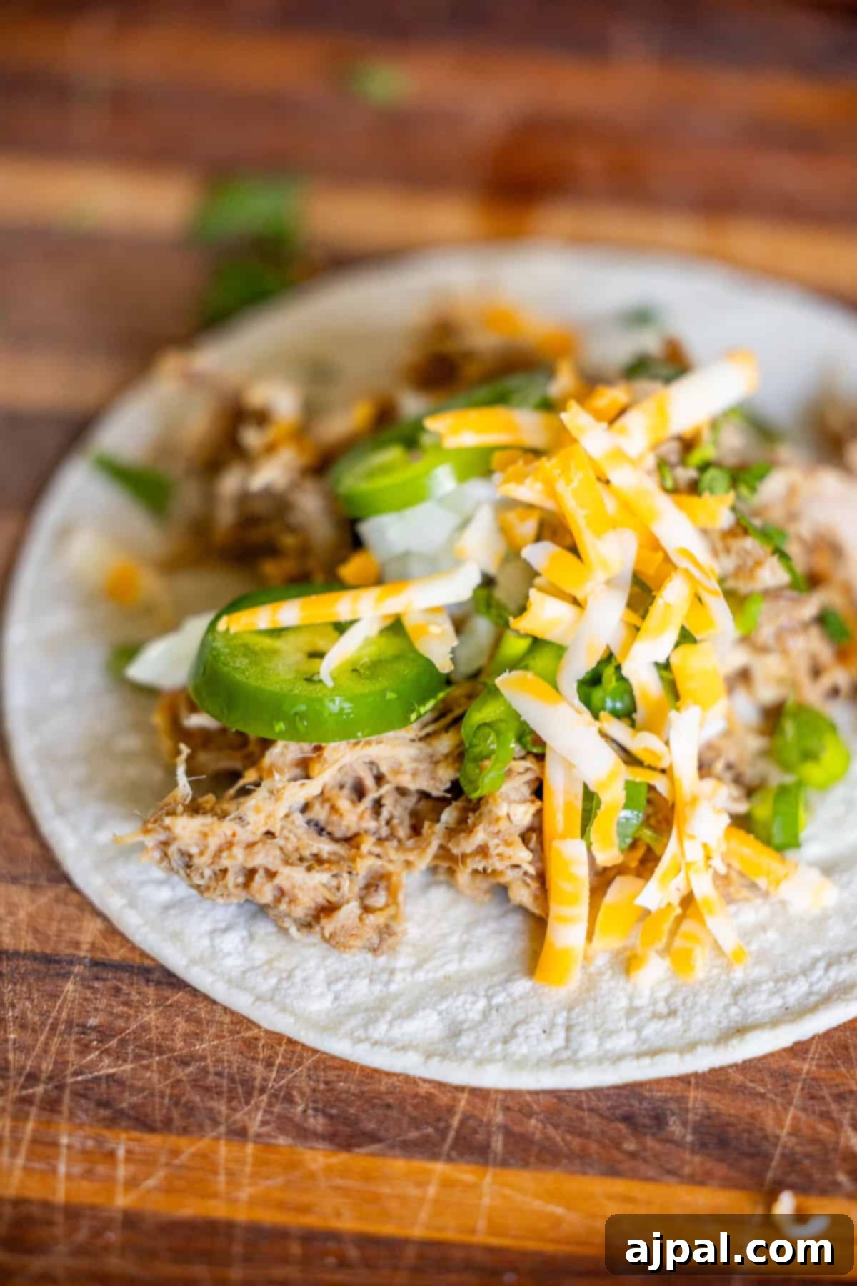 A flour tortilla topped with Slow Cooker Carnitas, sliced jalapeños, chopped onions, and shredded cheese on a wooden surface.