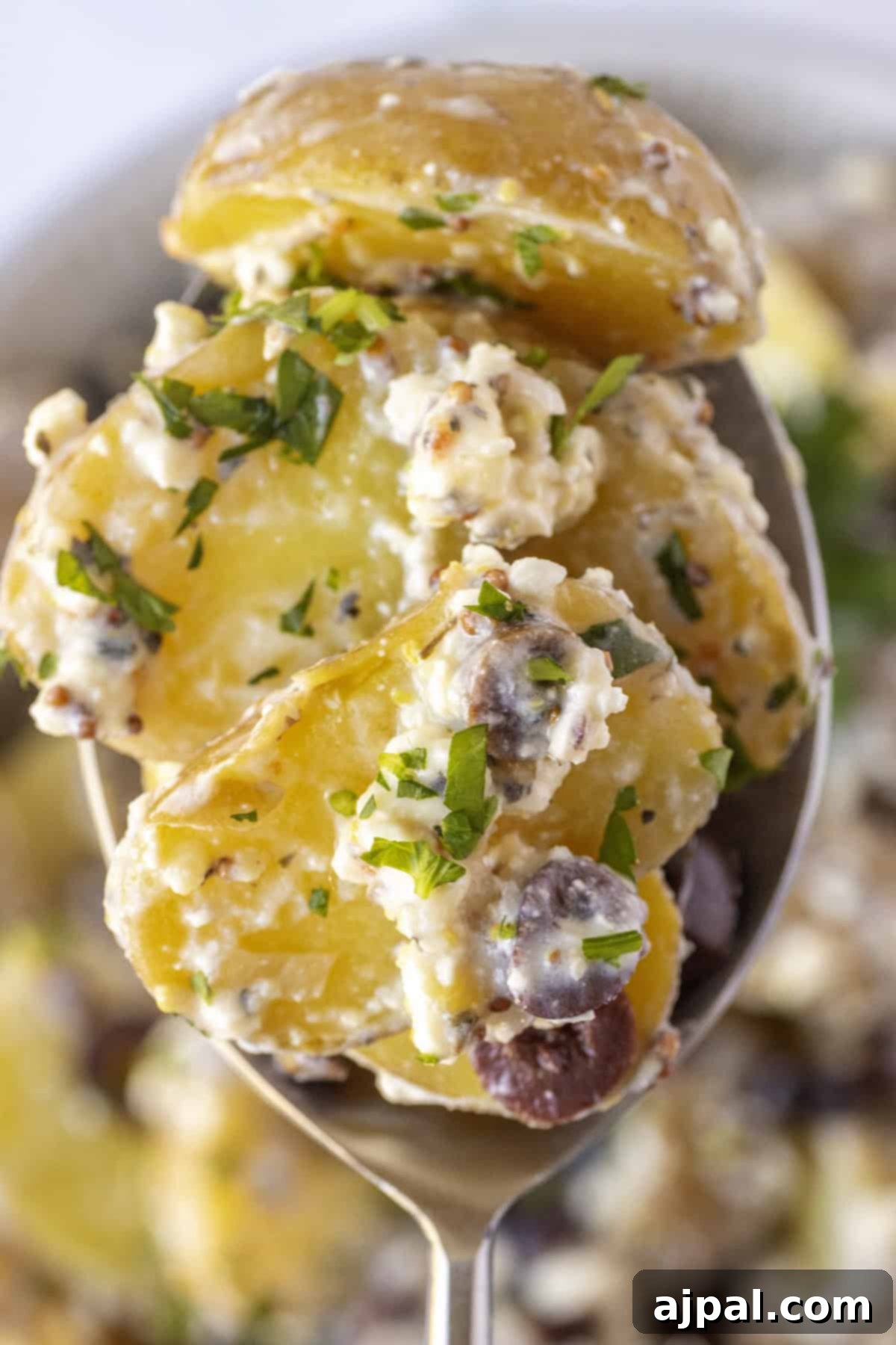 Classic Greek Potato Salad 2 Close-up of a spoonful of Greek potato salad with chopped parsley, sliced tender potatoes, creamy dressing, and small pieces of Kalamata olives.