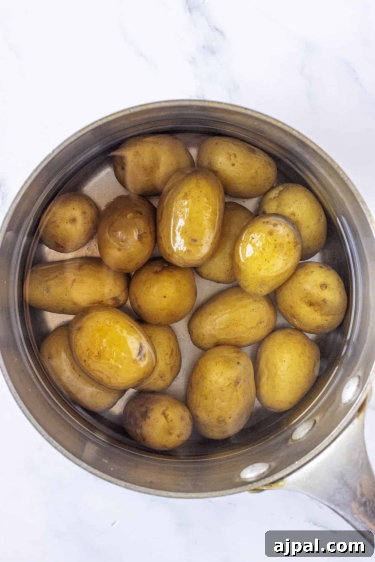 Classic Greek Potato Salad 3 A stainless steel pot filled with whole, unpeeled baby potatoes submerged in water on a white marble surface, ready to be cooked for Greek Potato Salad.