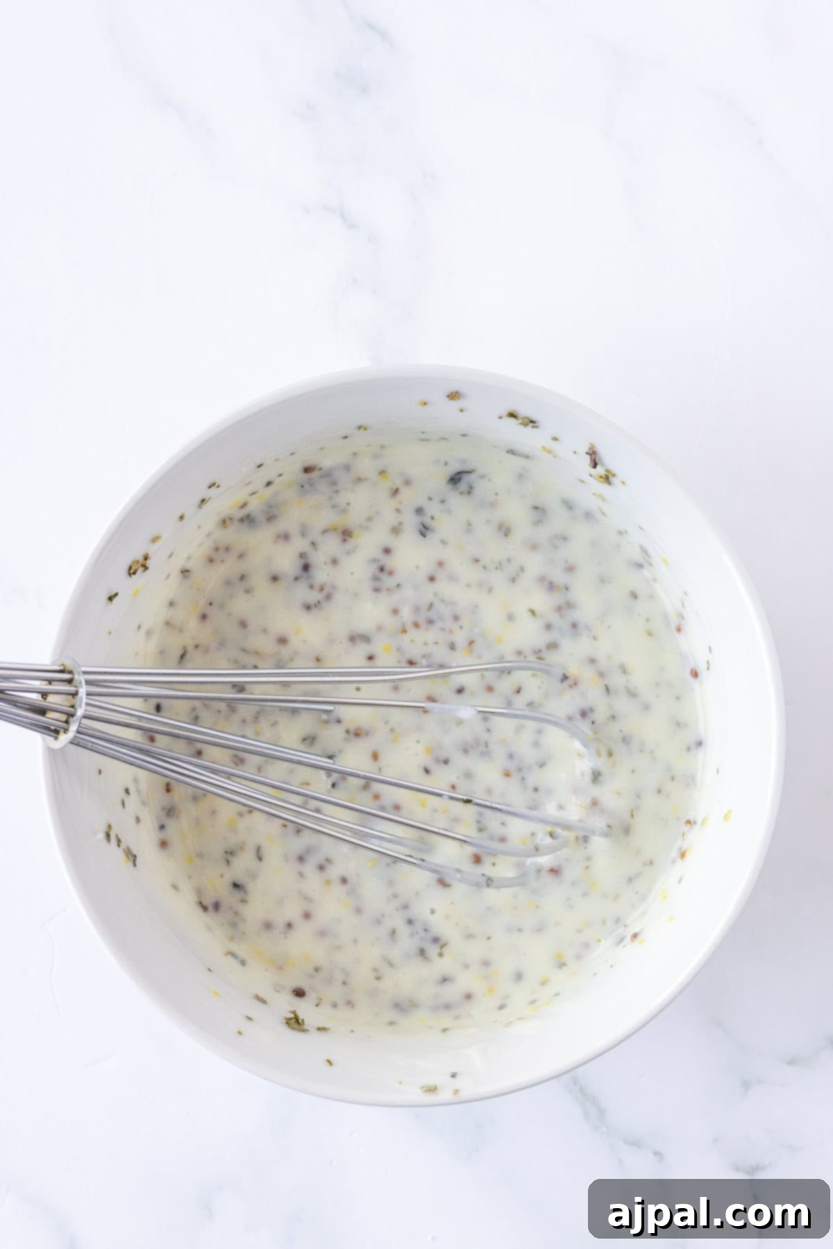 Classic Greek Potato Salad 4 A metal whisk in a white bowl filled with creamy, speckled Greek Potato Salad dressing or sauce rests on a white marble surface, demonstrating the vinaigrette preparation.