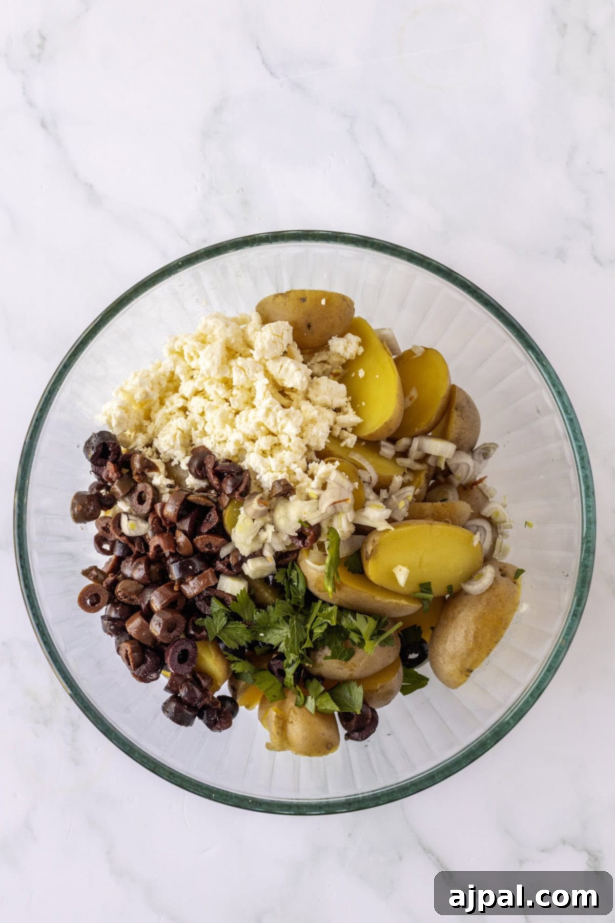 Classic Greek Potato Salad 5 Glass bowl with sliced yellow potatoes, chopped black olives, crumbled feta cheese, finely diced shallots, and fresh parsley on a white marble surface, ready for mixing to create a vibrant Greek Potato Salad.