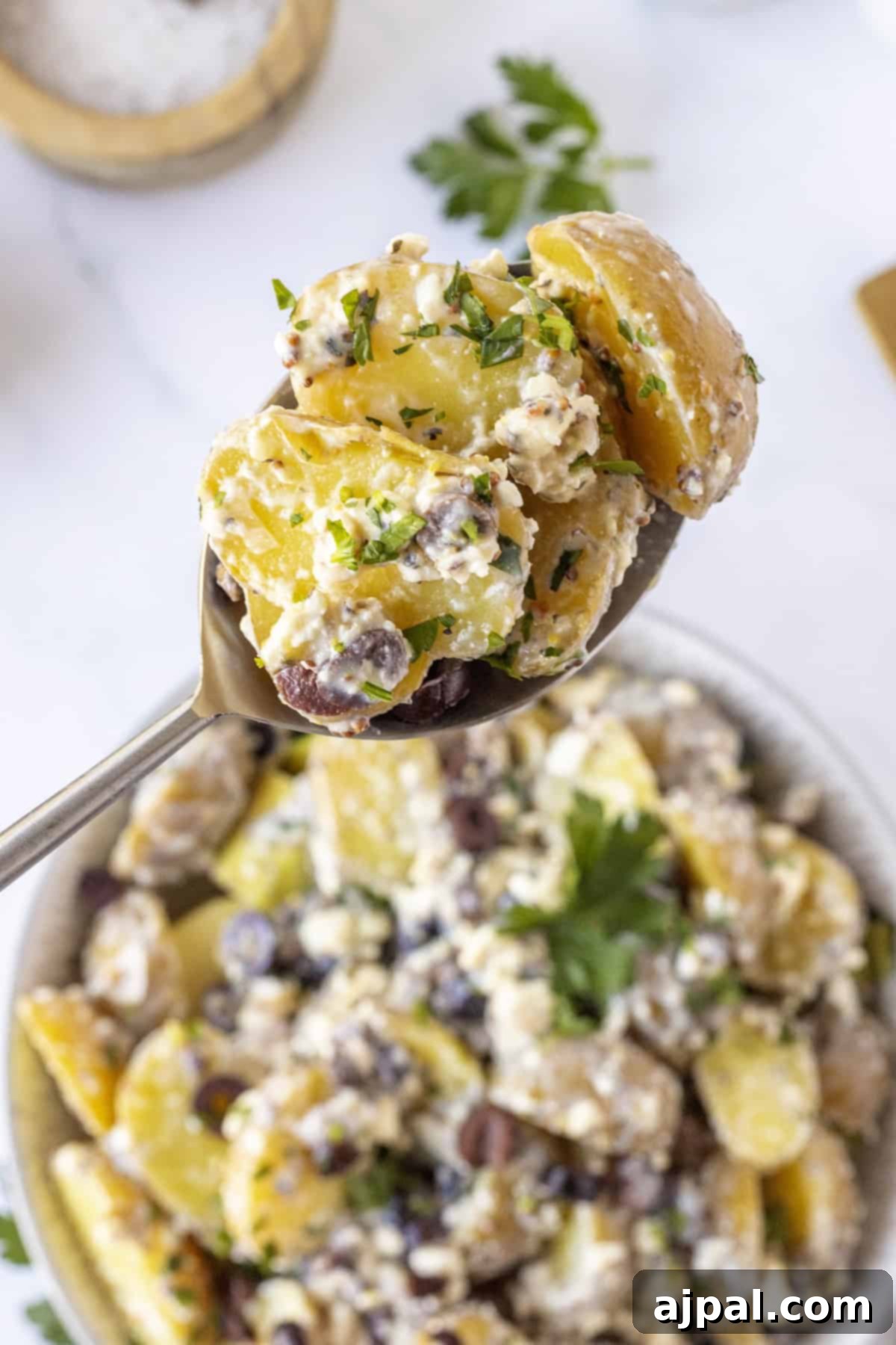 Classic Greek Potato Salad 10 A spoonful of Greek Potato Salad with herbs, olives, and creamy dressing is held above a bowl of the same salad on a white surface, showcasing its texture.