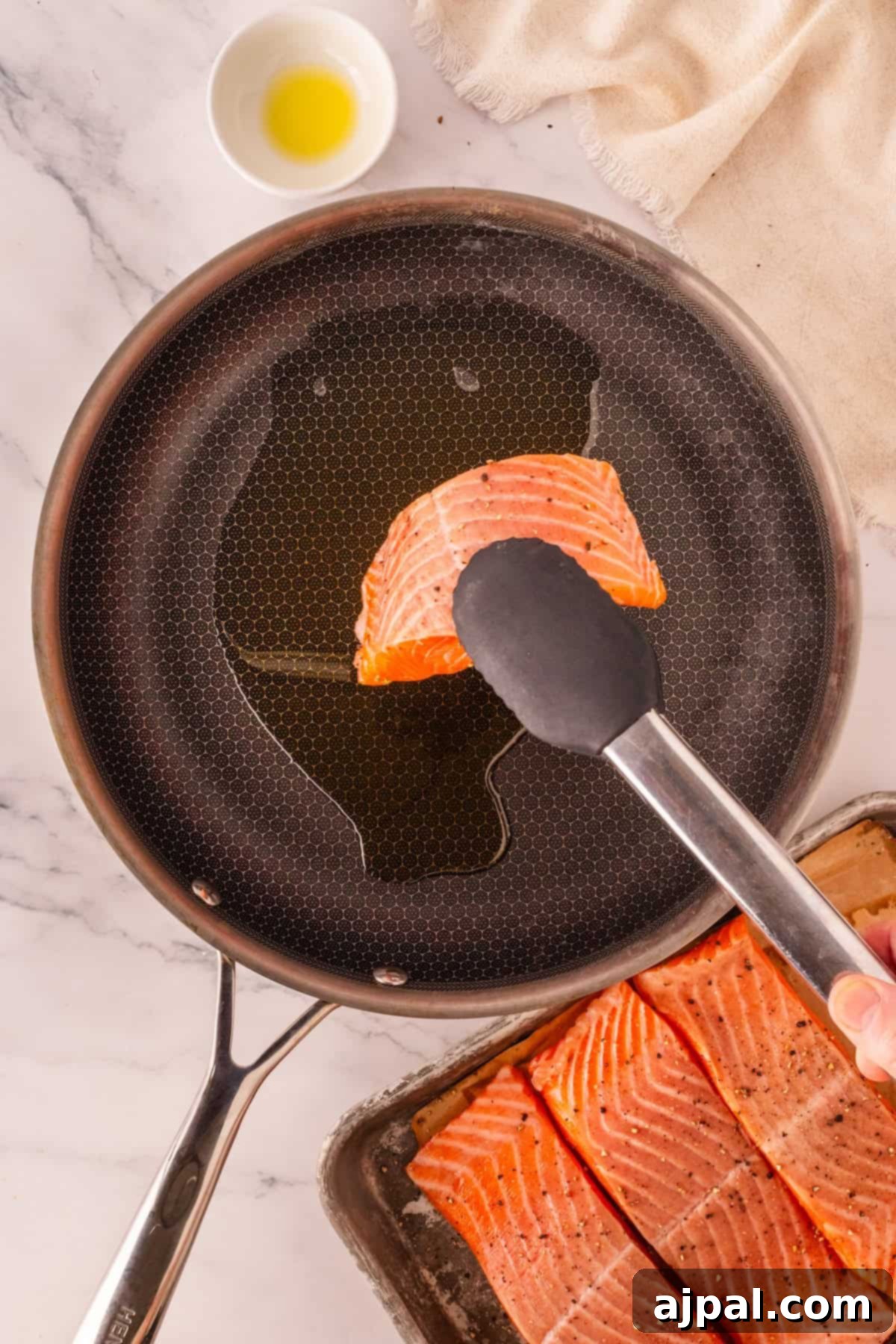 A chef expertly uses tongs to place a fresh raw salmon fillet into a sizzling pan of olive oil on a pristine marble countertop, skillfully preparing delicious Garlic Butter Salmon. More fillets and a small bowl of oil await nearby.