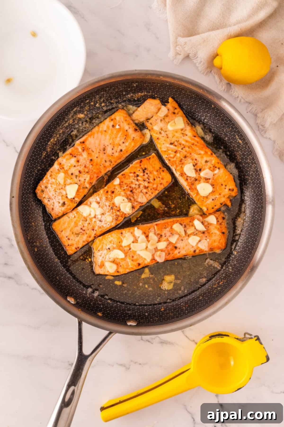 Four salmon fillets with garlic slices are cooking in a skillet, featuring the best baked salmon recipe, with a lemon, juicer, and white plate nearby on a marble countertop.