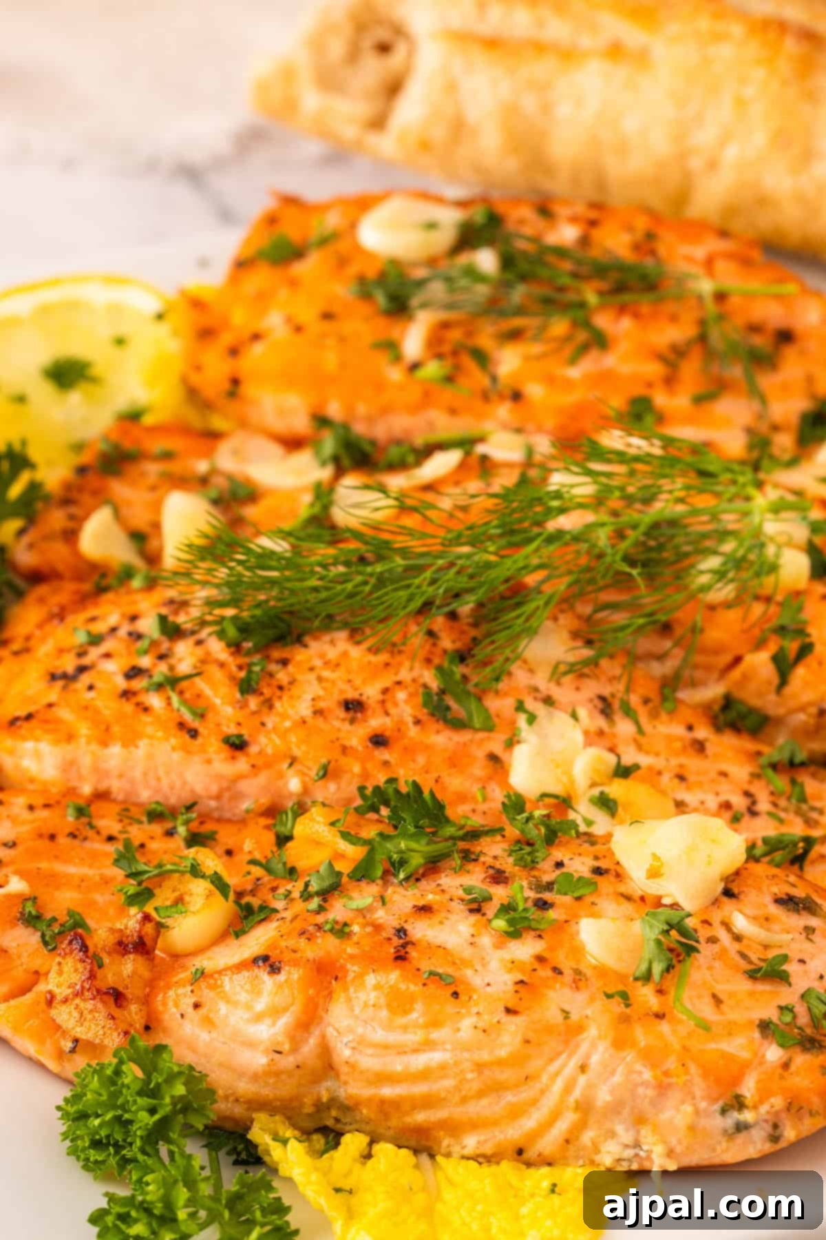 Grilled garlic butter salmon fillets garnished with fresh dill, parsley, lemon slices, and sliced garlic, with a piece of bread in the background.