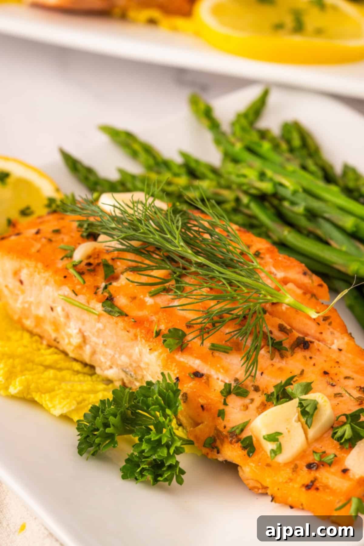 A succulent Garlic Butter Salmon fillet, artfully garnished with fresh herbs and bright lemon slices, perfectly served alongside tender asparagus spears on a pristine white plate, creating a balanced and flavorful meal.