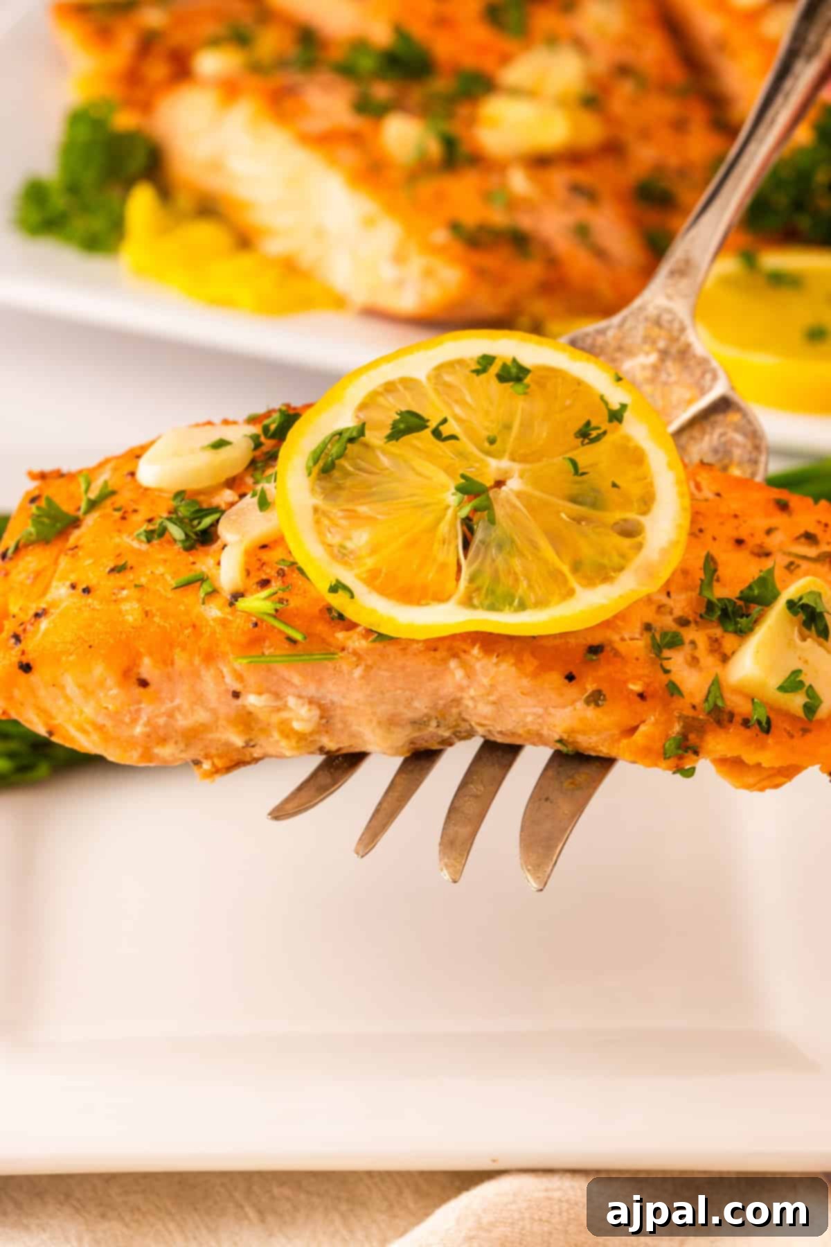A fork elegantly holds a perfectly cooked Garlic Butter Salmon fillet, beautifully topped with fresh lemon slices, aromatic herbs, and savory garlic, with more salmon and vibrant leafy greens artfully arranged in the background, ready to be enjoyed.