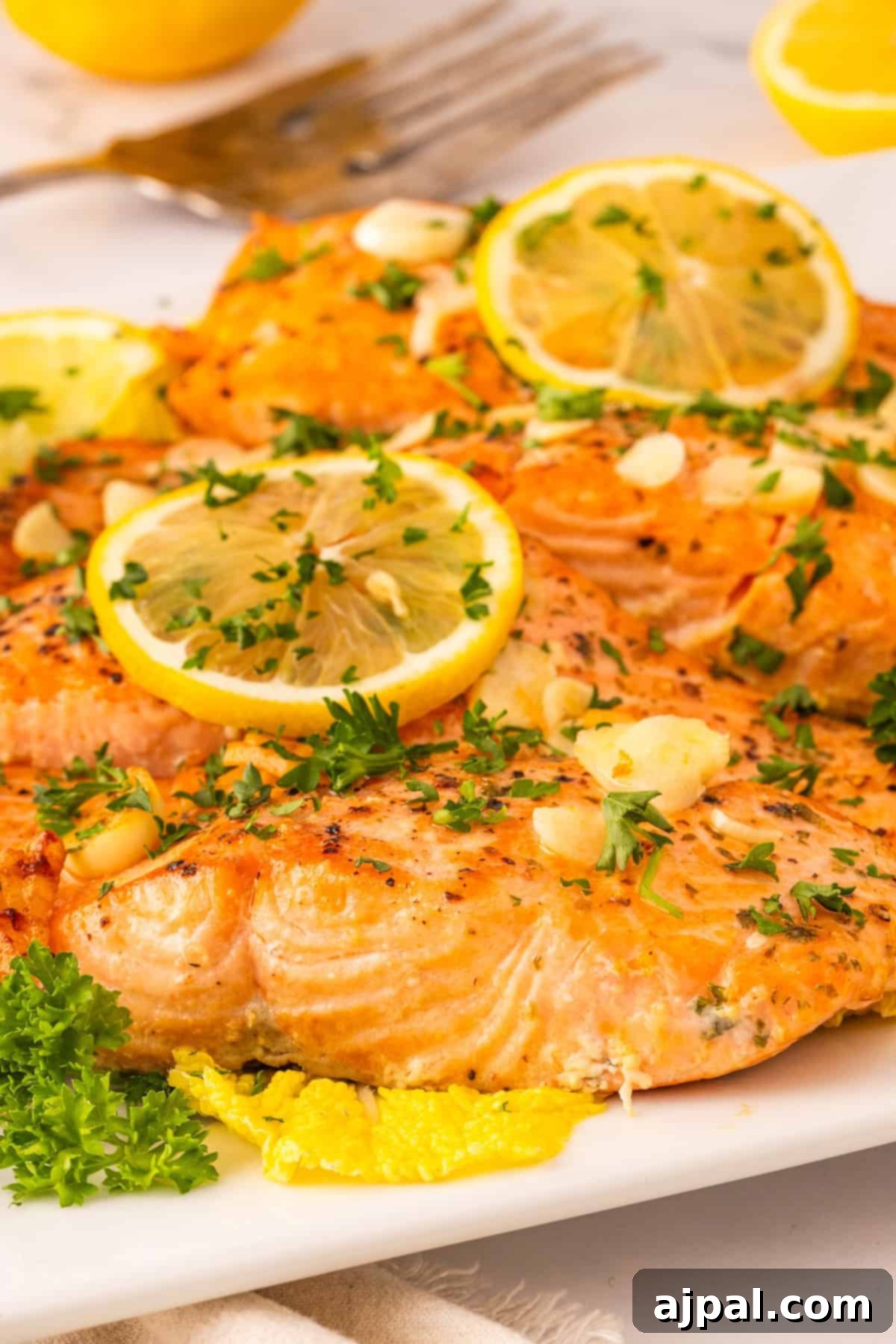 Exquisitely arranged garlic butter salmon fillets, adorned with bright lemon slices, finely chopped parsley, and savory garlic, presented on a pristine white plate with additional lemon wedges and a fork in the background, ready for a delightful meal.