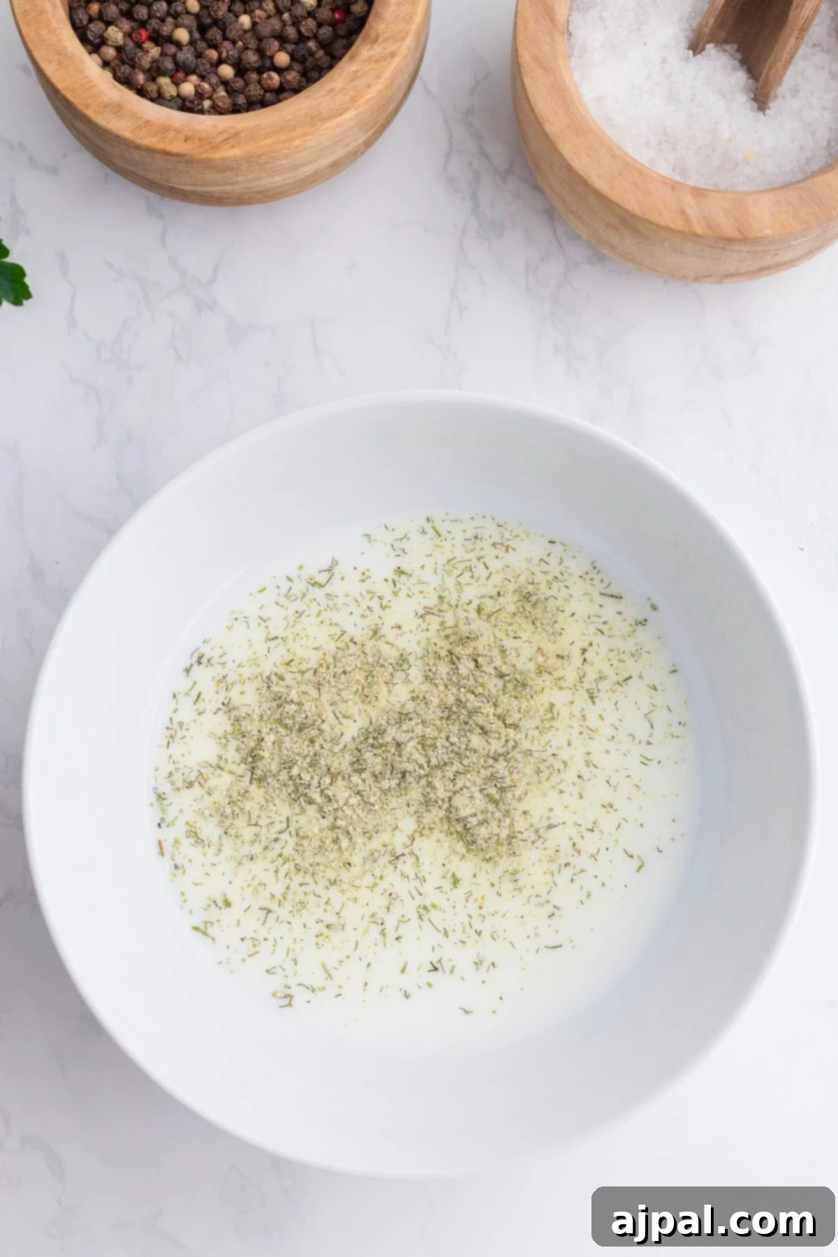 Ranch Infused Chicken Breasts 3 A white bowl with a creamy liquid and dried herbs, perfect for Ranch Chicken Breasts, placed on a marble surface next to bowls of peppercorns and salt.