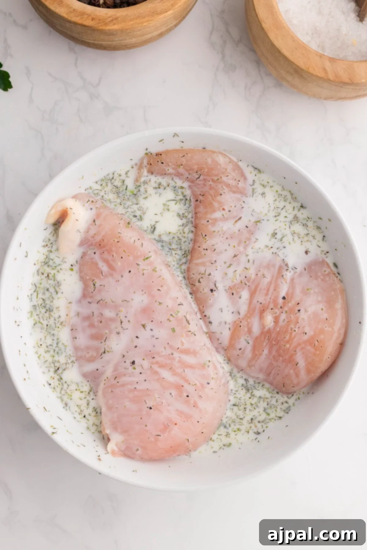 Ranch Infused Chicken Breasts 4 Raw Ranch Chicken Breasts marinating in a white bowl with herbs and a creamy liquid, set on a white marble surface beside wooden bowls of salt and spices.