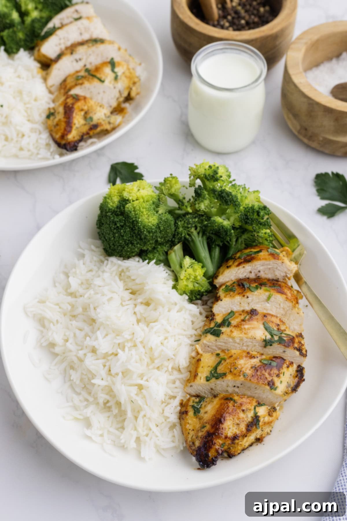 Ranch Infused Chicken Breasts 9 A plate with sliced Ranch Chicken Breasts, steamed broccoli, and white rice, served with a fork. A small jar of sauce and bowls of pepper and salt are in the background.