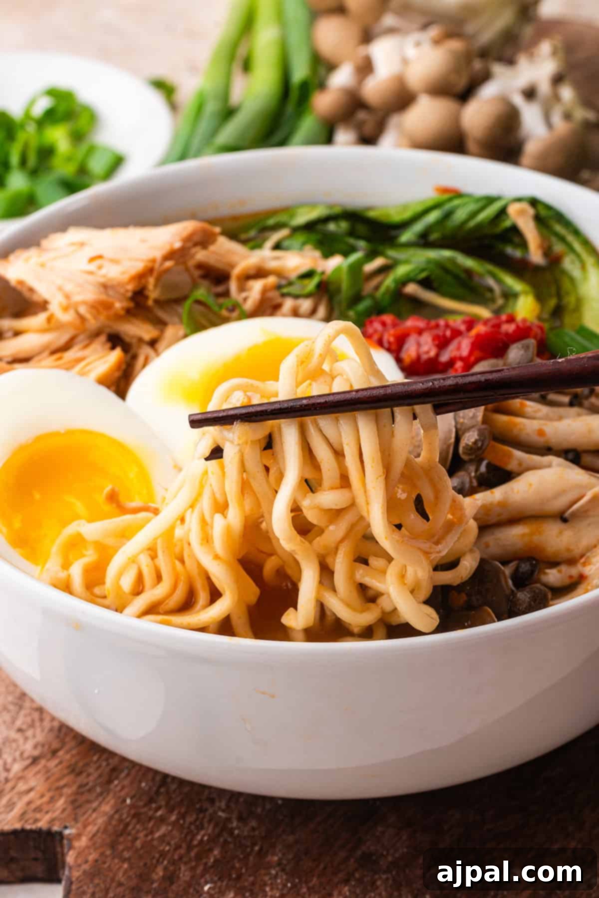 A bowl of Chicken Ramen features noodles, soft-boiled egg, mushrooms, shredded slow cooker chicken, fresh greens, and chopsticks lifting noodles for a comforting meal.