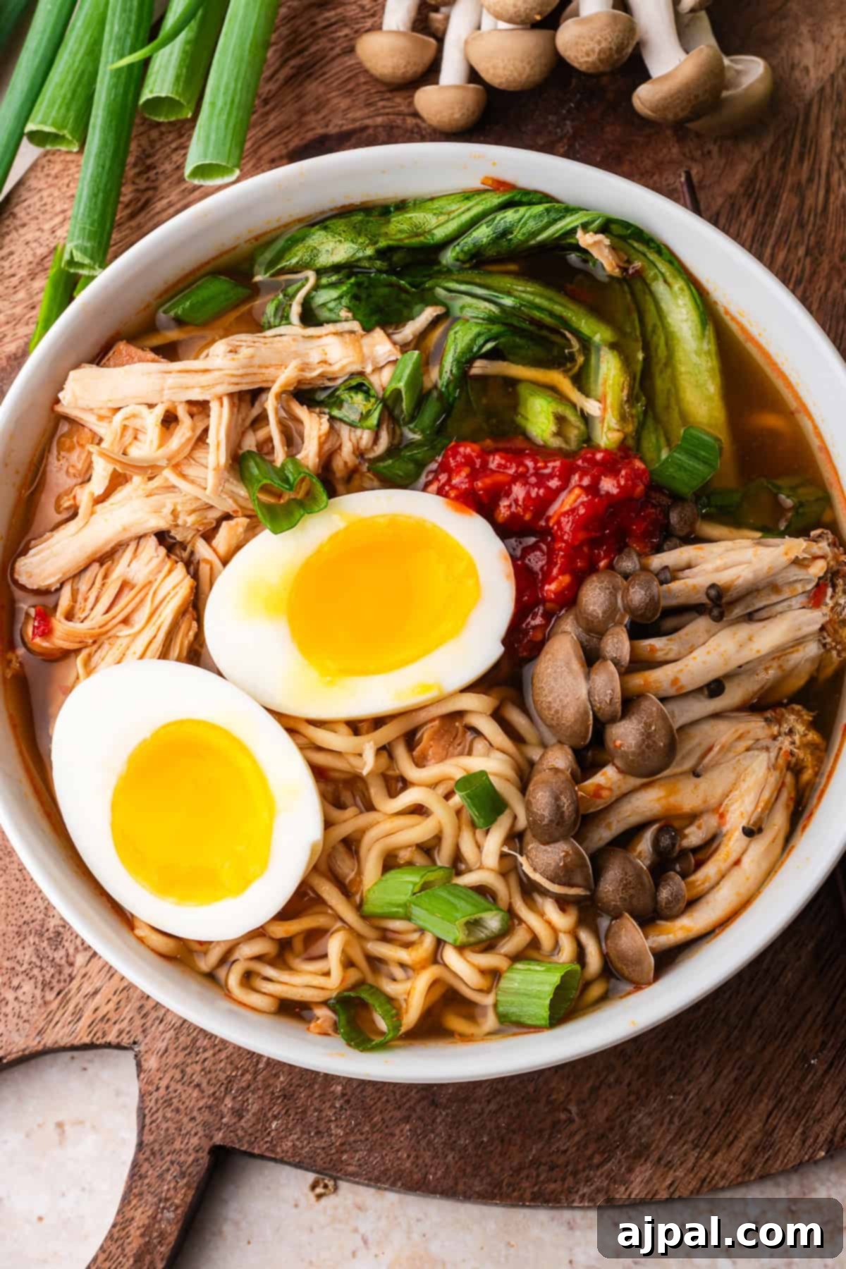 A bowl of Slow Cooker Chicken Ramen topped with soft-boiled eggs, shredded chicken, mushrooms, greens, scallions, and chili paste sits on a wooden board with green onions and mushrooms nearby.