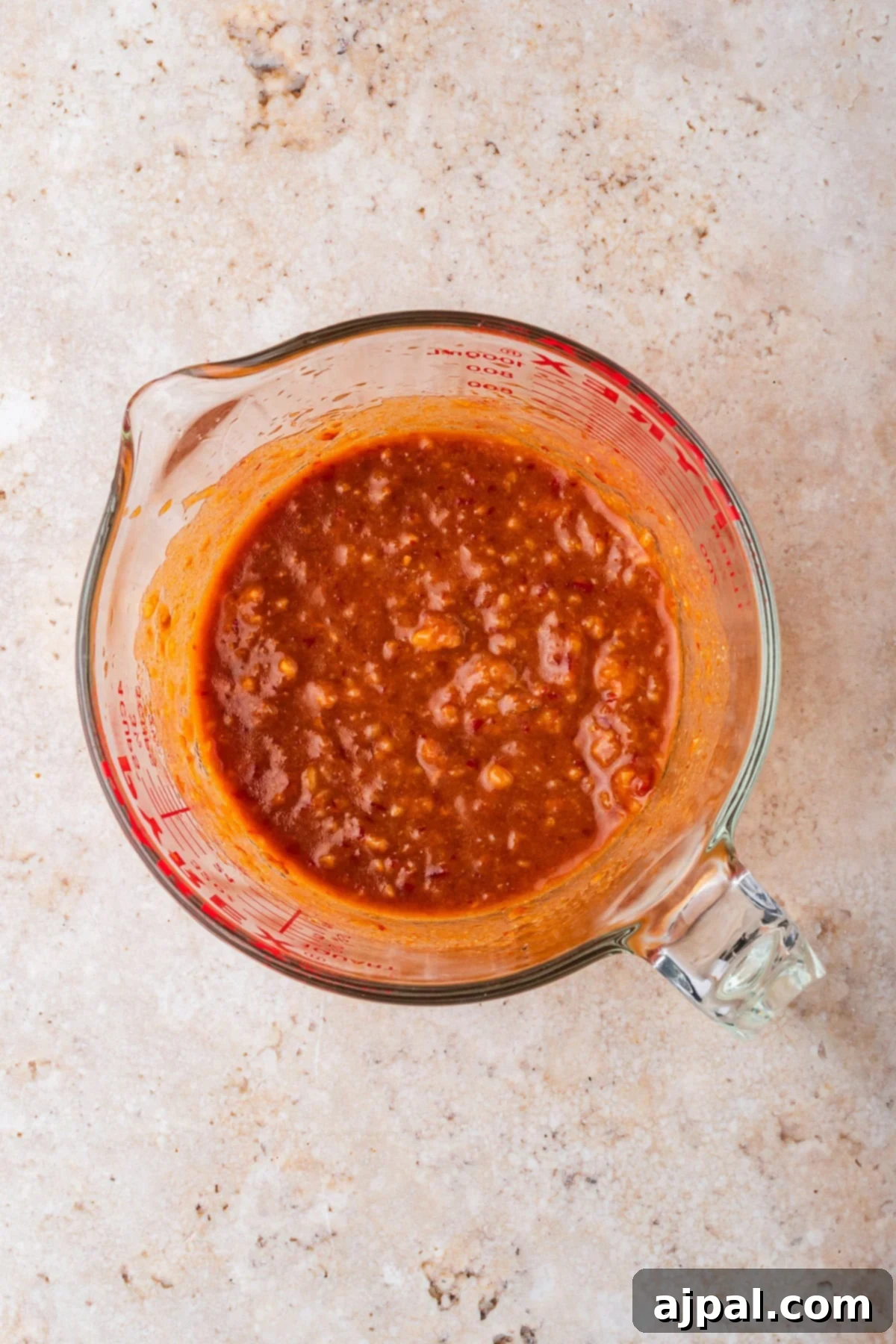 Glass measuring cup filled with chunky red sauce or salsa, perfect for topping your favorite Chicken Ramen or adding extra flavor to Slow Cooker recipes. Placed on a light-colored, textured surface.