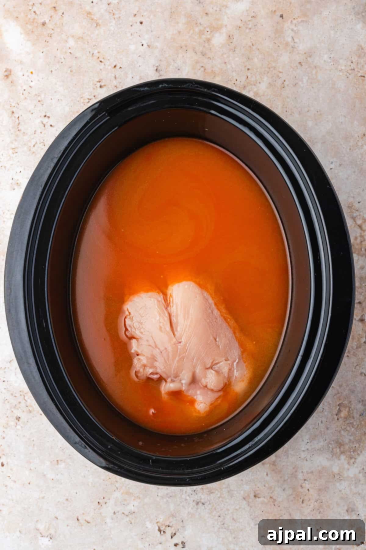 A raw chicken breast rests in orange-colored liquid inside a black slow cooker, perfect for preparing flavorful Slow Cooker Ramen, all set on a light brown countertop.