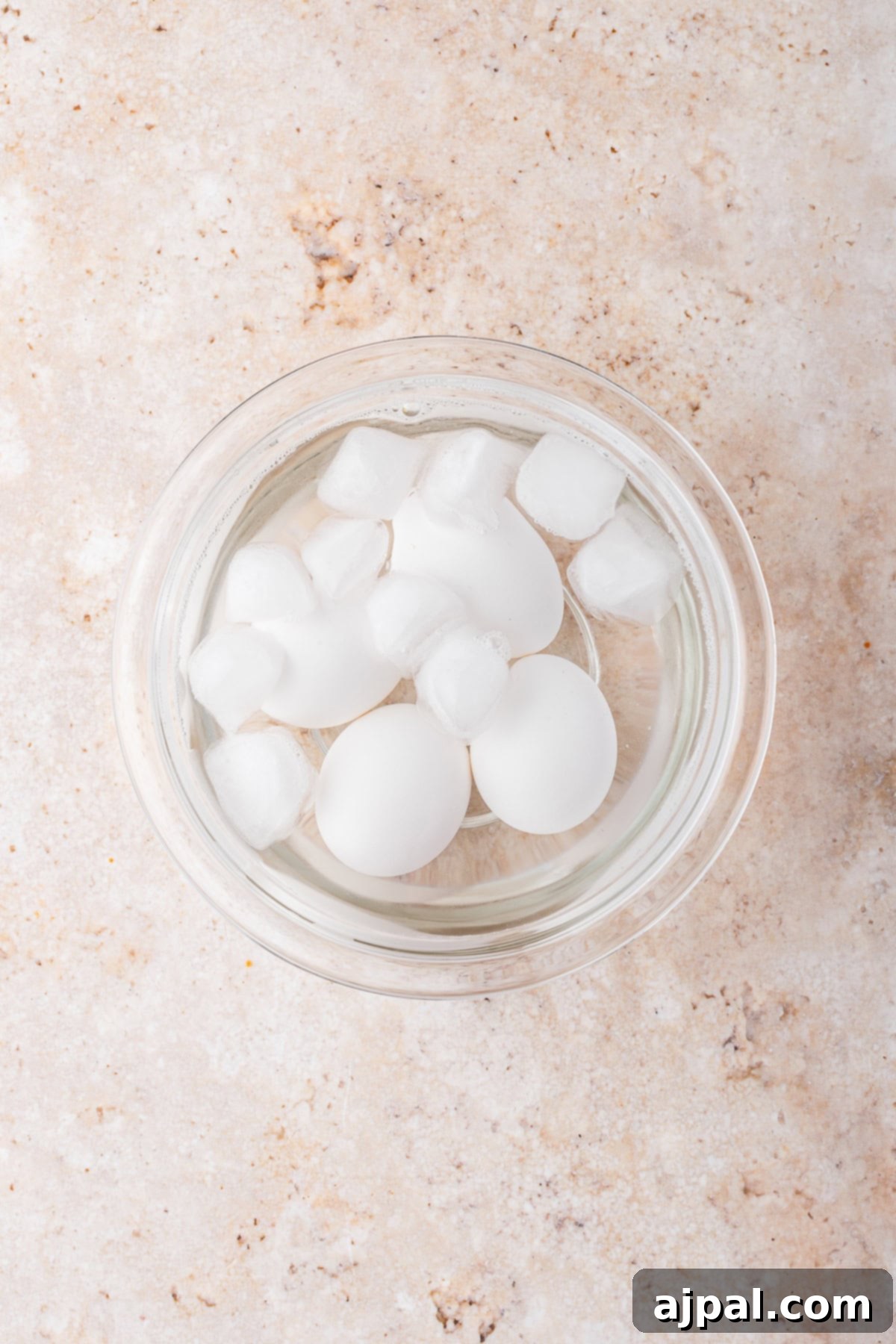A glass bowl filled with water, ice cubes, and three white eggs sits on a light-colored, textured surface—perfect for prepping ingredients for your favorite Slow Cooker Chicken Ramen.