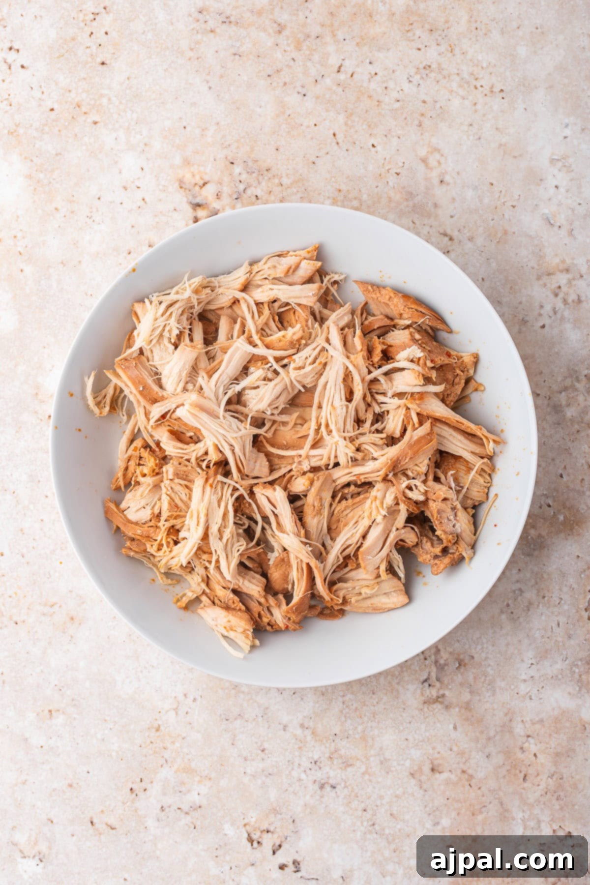 A white bowl filled with shredded slow cooker chicken sits on a light-colored, textured surface—perfect for adding to your favorite chicken ramen.