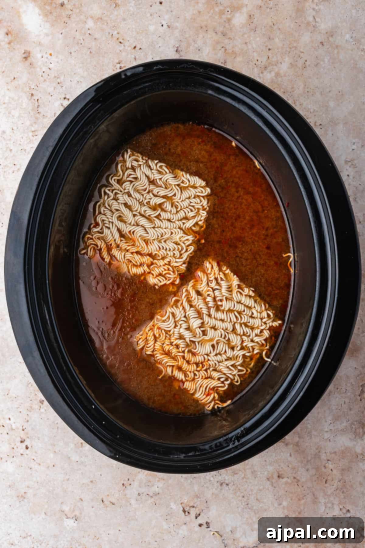 Two blocks of uncooked instant ramen noodles simmer in a slow cooker with savory broth, creating an easy and comforting Chicken Ramen meal.