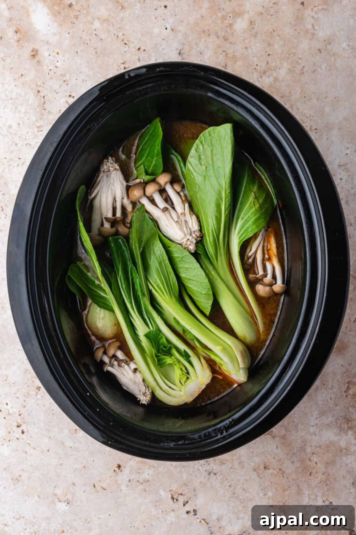 A slow cooker filled with bok choy, mushrooms, and broth for Slow Cooker Chicken Ramen, viewed from above on a light stone surface.