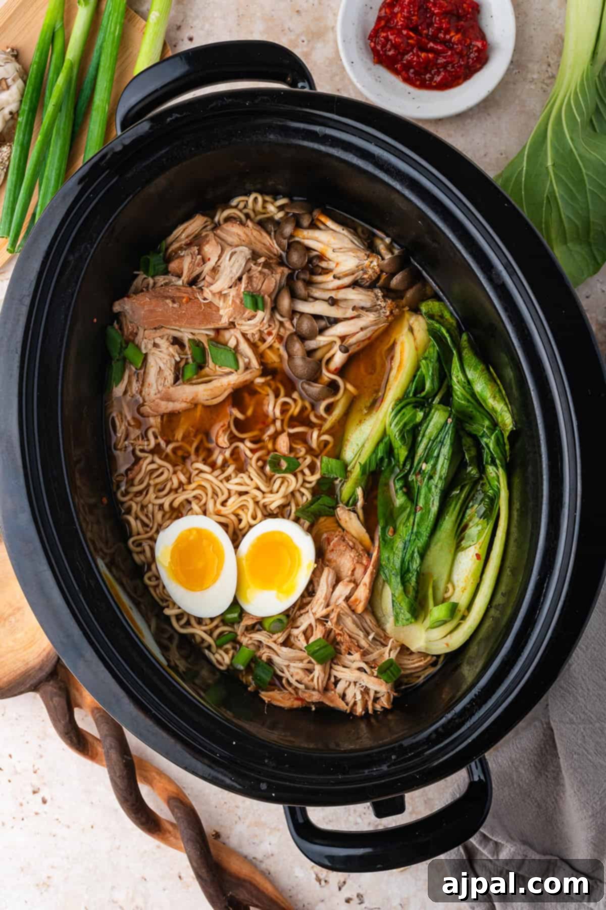 A bowl of Slow Cooker Ramen in a black pot with noodles, shredded chicken, soft-boiled eggs, mushrooms, and bok choy, garnished with green onions.