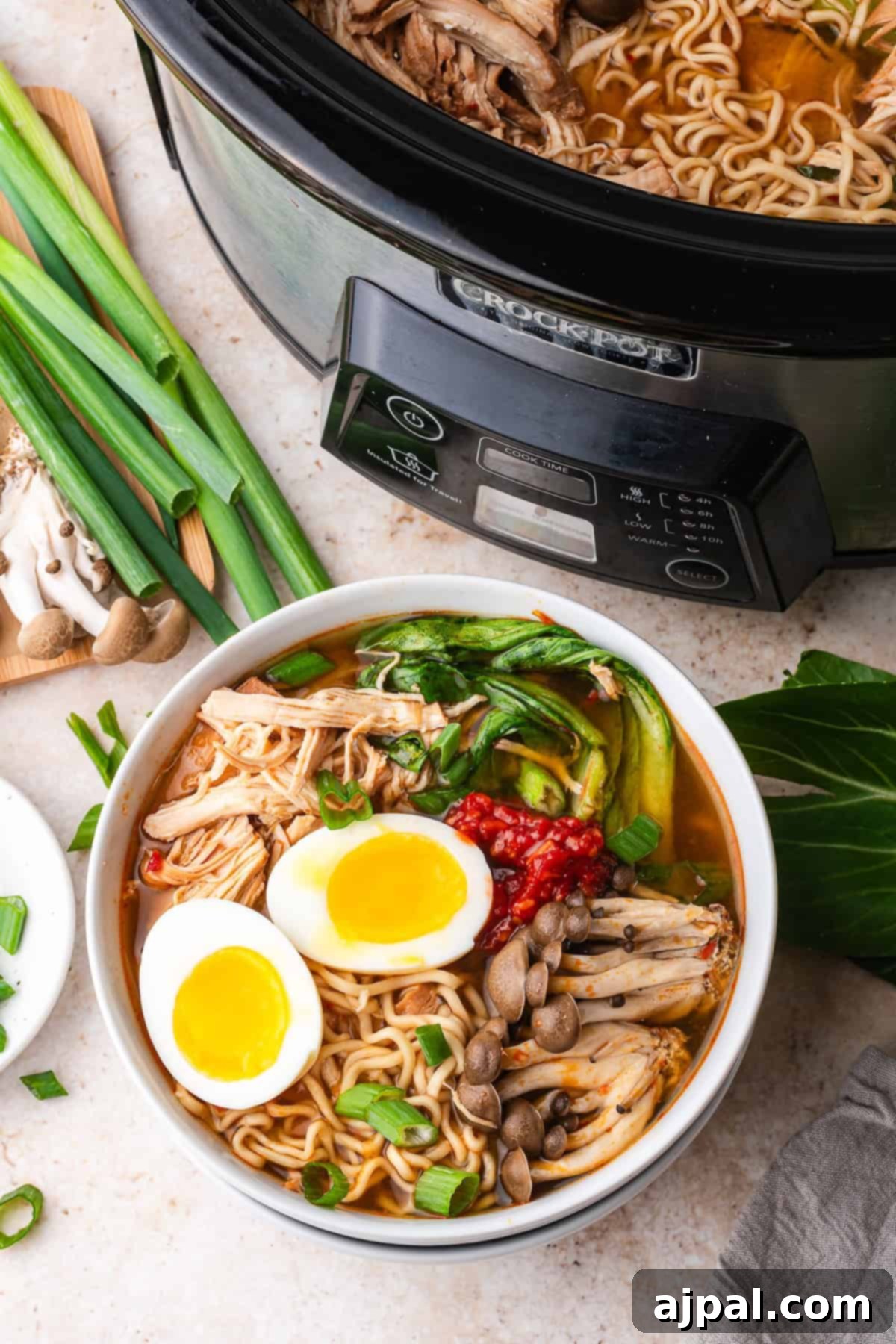 A bowl of Chicken Ramen with soft-boiled eggs, shredded chicken, mushrooms, greens, chili paste, and green onions sits next to a Slow Cooker filled with savory broth and tender chicken.