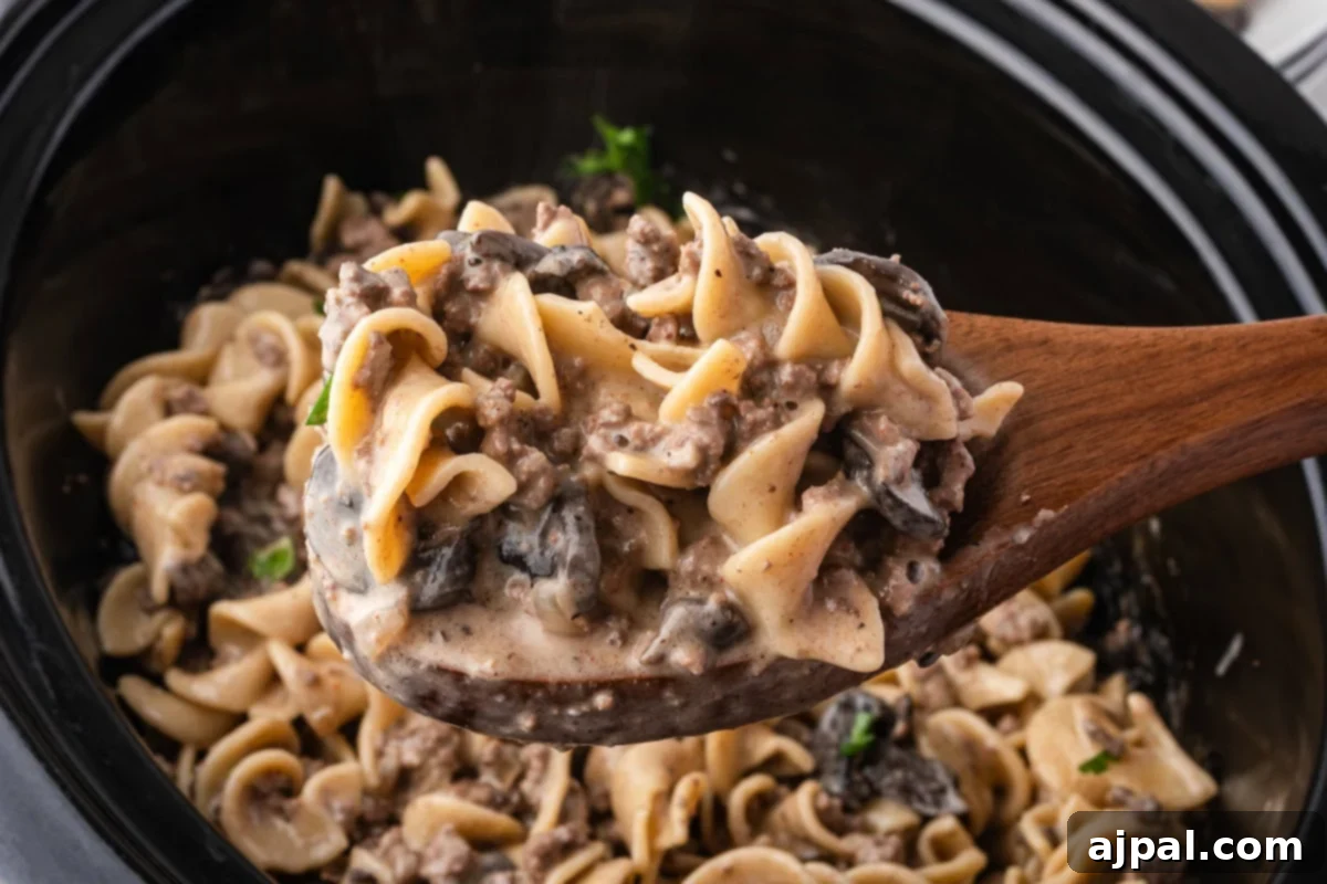 A wooden spoon holds a portion of creamy Slow Cooker Beef Stroganoff with egg noodles, mushrooms, and ground beef, above a slow cooker brimming with the savory dish.