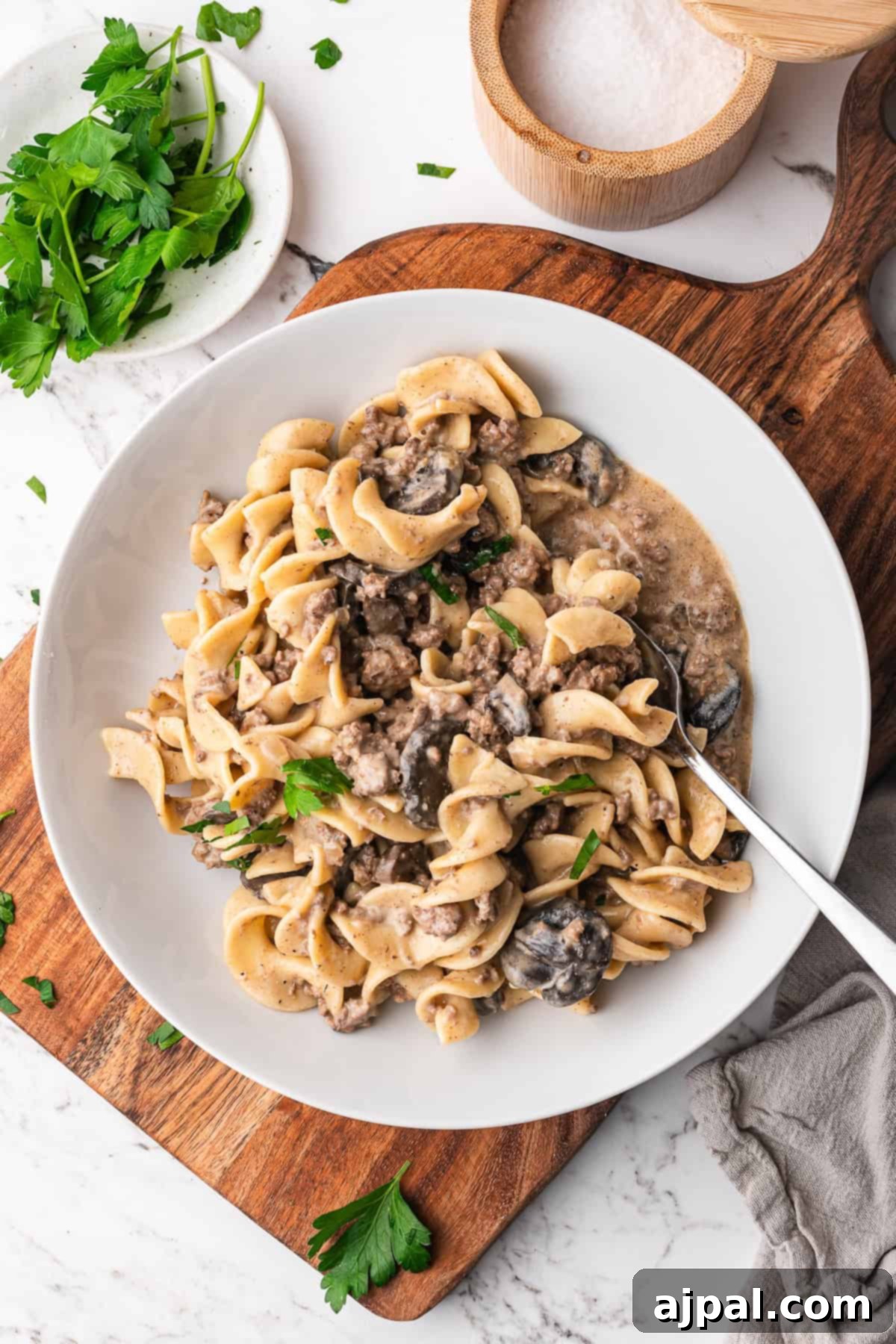 A bowl of Slow Cooker Beef Stroganoff with egg noodles and mushrooms, garnished with parsley, sits on a wooden board with a fork, parsley, a salt cellar, and a napkin nearby.