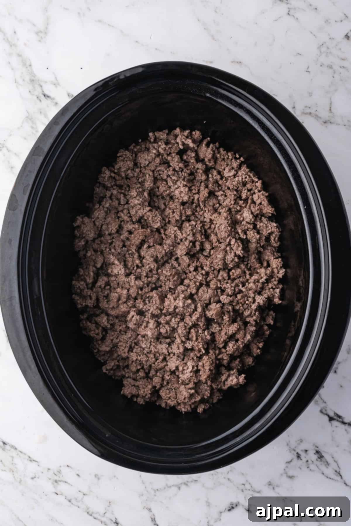Tender cooked ground beef in a black slow cooker sits on a white marble surface—perfect for adding to your favorite beef recipes like classic Beef Stroganoff.