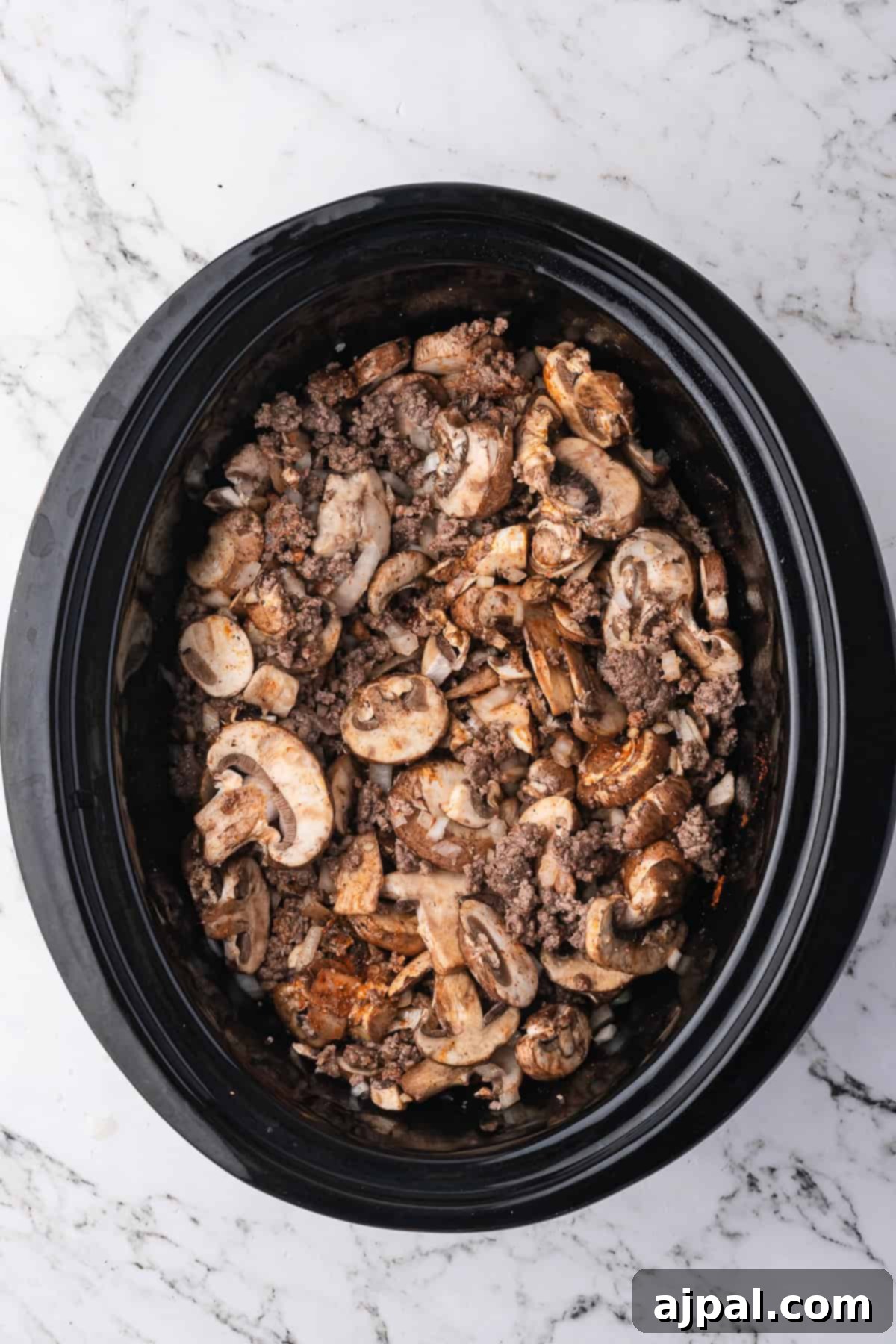 A black slow cooker filled with sliced mushrooms and ground beef sits on a white marble surface, perfect for making classic Beef Stroganoff or other hearty beef recipes.