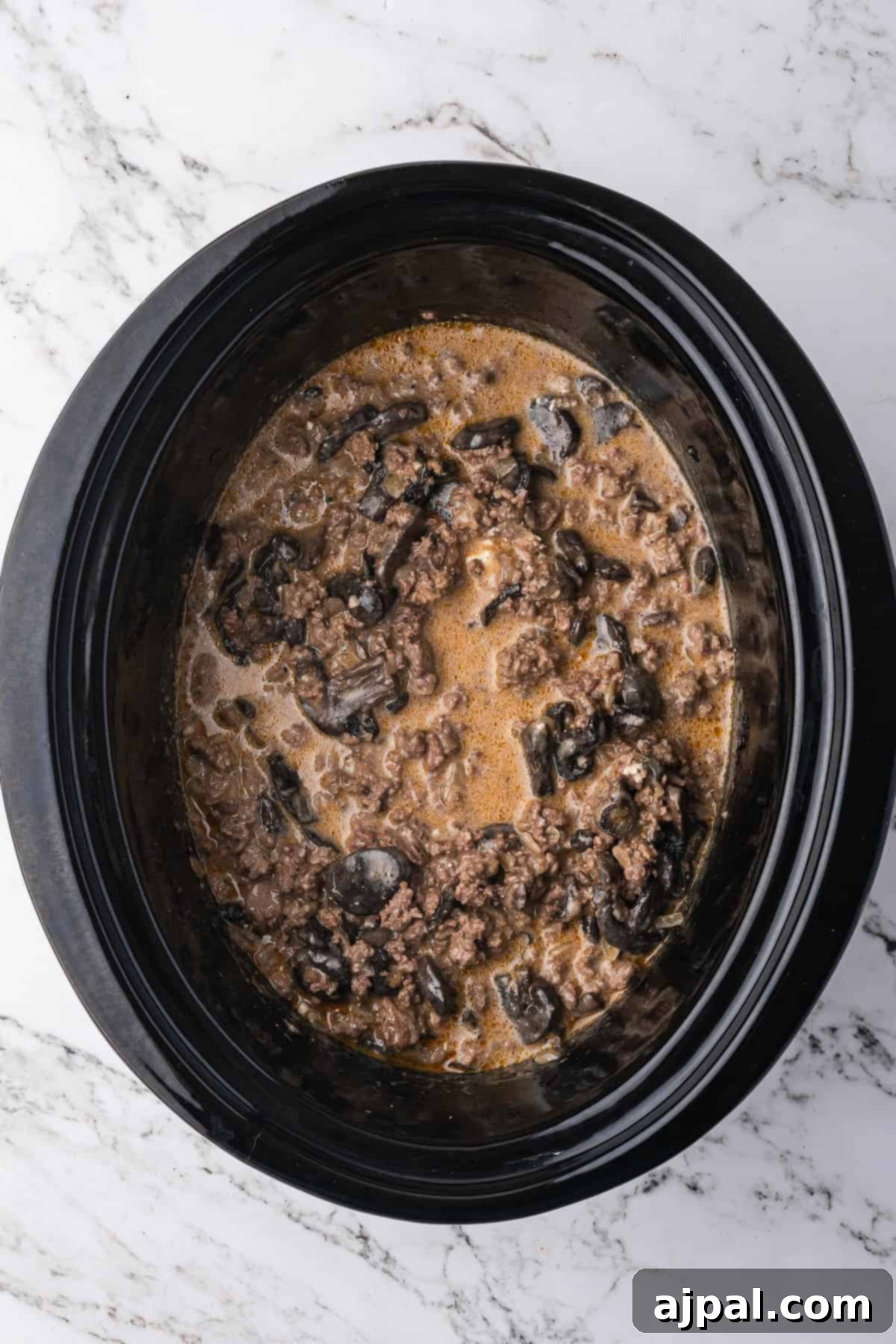 Ground beef, black olives, and a savory sauce mixture simmer together in a black slow cooker, perfect for creating an easy Slow Cooker Beef Stroganoff on a marble countertop.