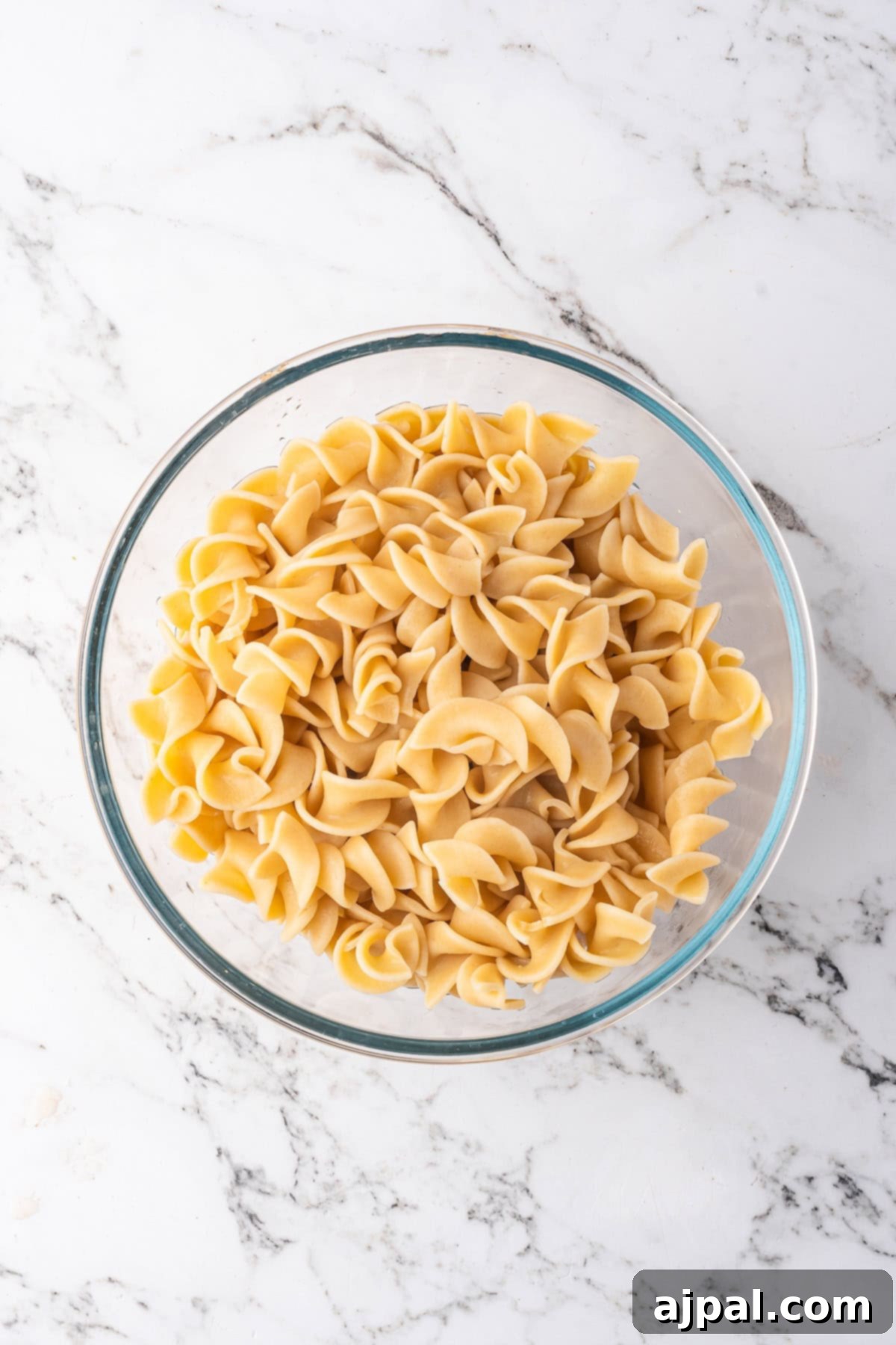A glass bowl filled with plain, cooked egg noodles sits on a white marble surface, ready to be topped with your favorite Beef Stroganoff or slow cooker beef recipe.