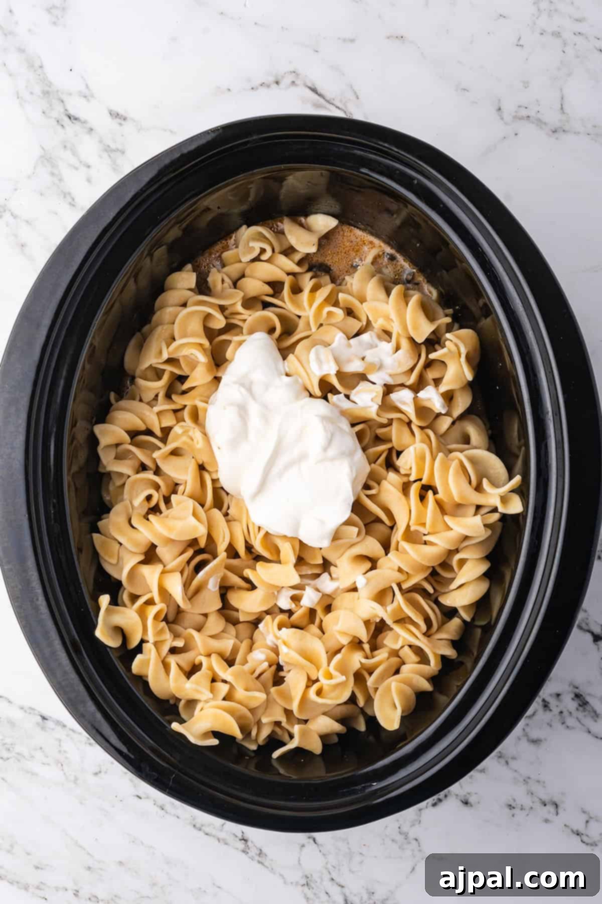 Uncooked egg noodles and a dollop of sour cream sit in a black slow cooker on a marble countertop, ready to become delicious Slow Cooker Beef Stroganoff.