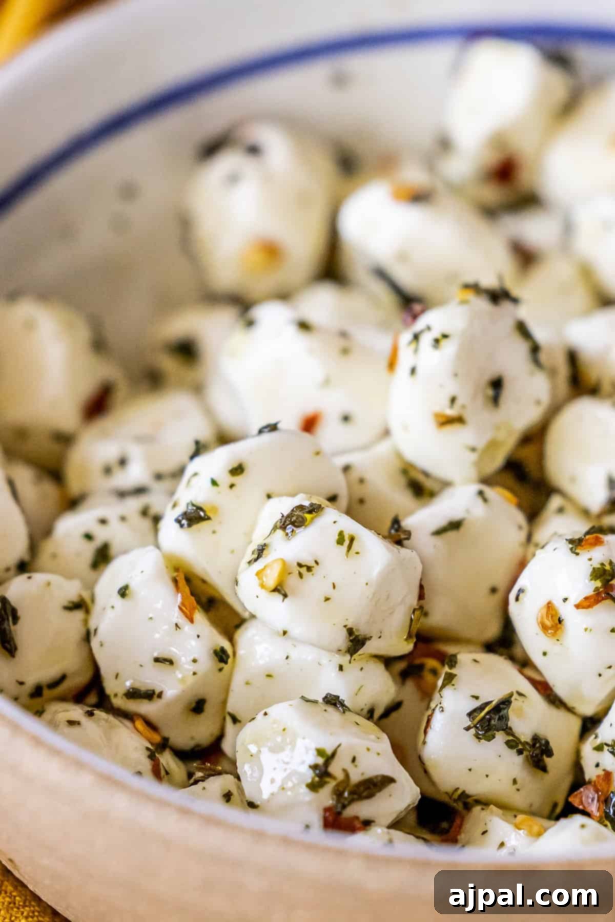 A close-up of perfectly marinated mozzarella balls coated with fresh herbs and spices, glistening in olive oil, served in an elegant ceramic bowl. The ideal appetizer for any occasion.