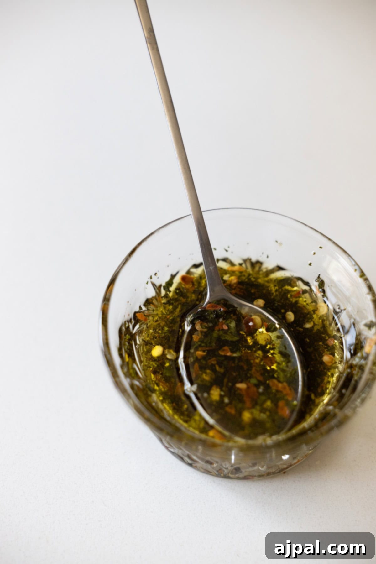 A glistening glass bowl filled with rich extra virgin olive oil, vibrant dried herbs, and fiery red pepper flakes, perfectly mixed for an authentic Marinated Mozzarella recipe on a pristine white surface.