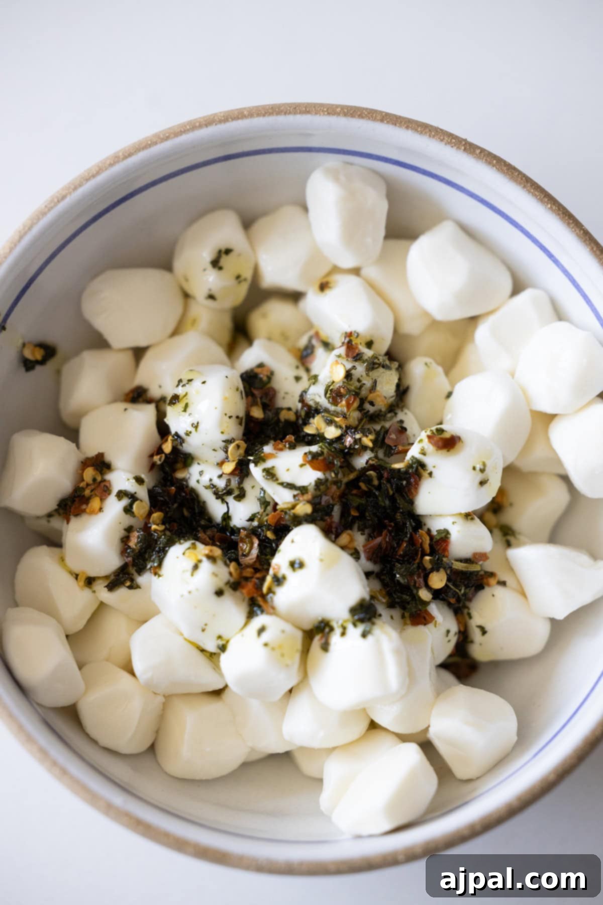 A bowl of marinated mozzarella balls, richly topped with a vibrant blend of dried herbs and red pepper flakes on a crisp white surface—a simple yet exquisitely flavorful mozzarella recipe ready to serve.